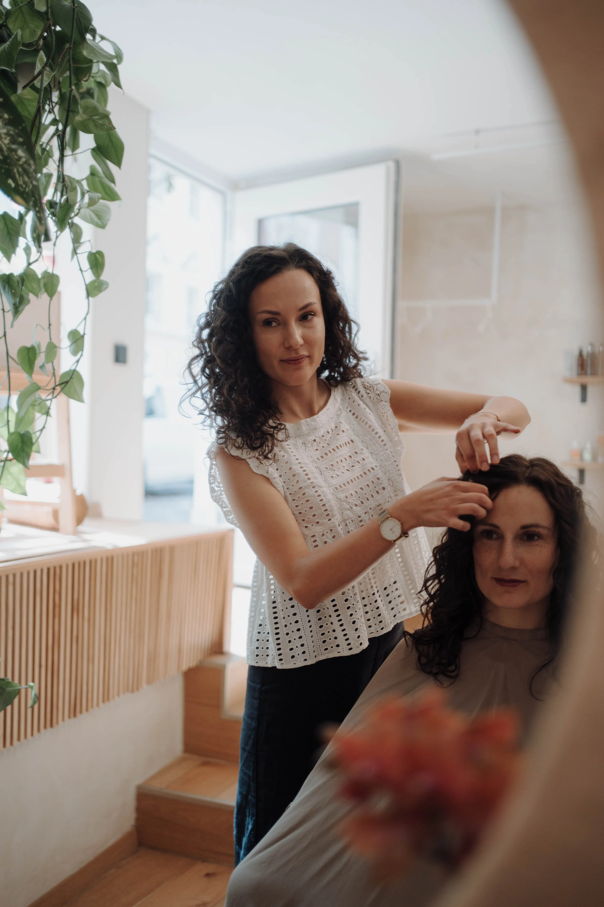Friseurin macht einer Kundin eine Frisur im gemütlichen Salon mit Pflanzen und großen Fenstern.