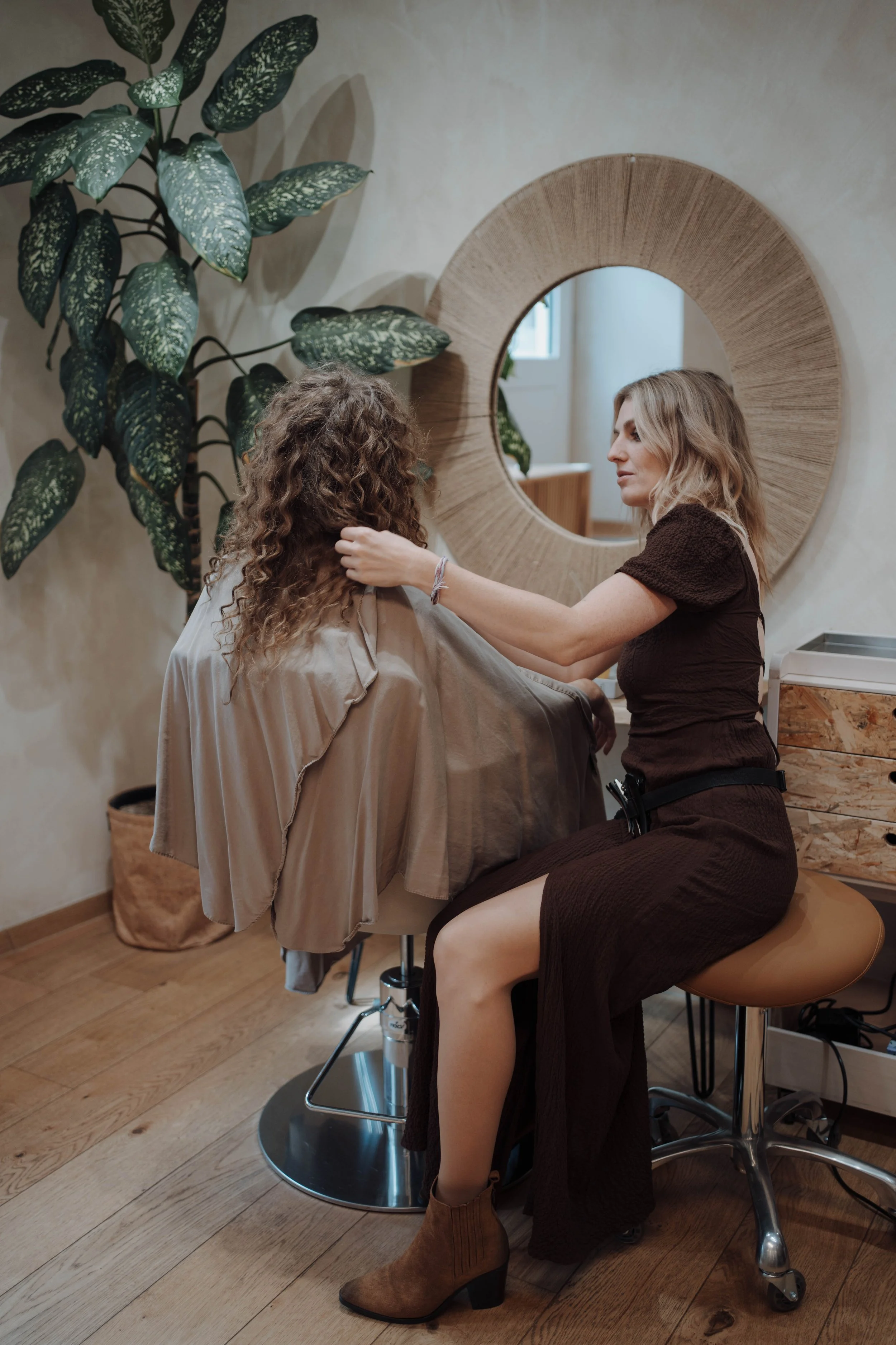 Friseurin schneidet die Haare einer Kundin in einem Friseursalon, mit großen Blättern und einem dekorativen runden Spiegel im Hintergrund.