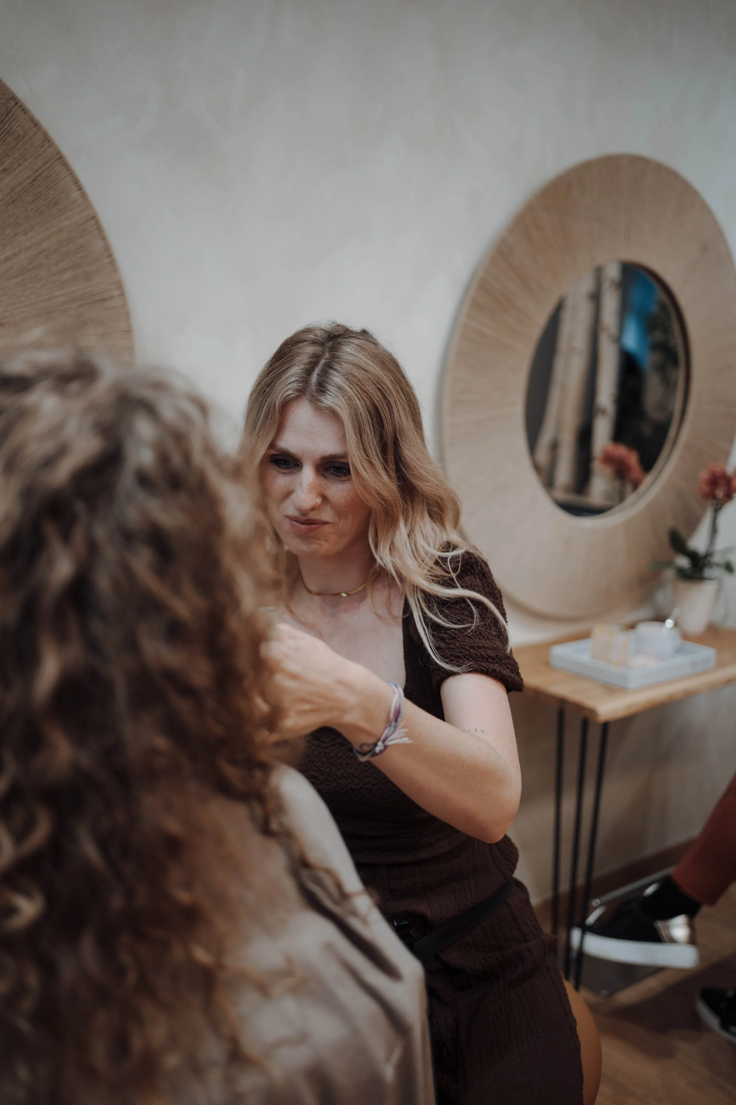 Eine Frau mit blonde Haare, die einen Perücken-Look trägt, wird von einer Stylistin frisiert. Sie sitzen in einem gemütlichen Raum mit runden Spiegeln an der Wand und einer kleinen Tisch mit Dekoration.