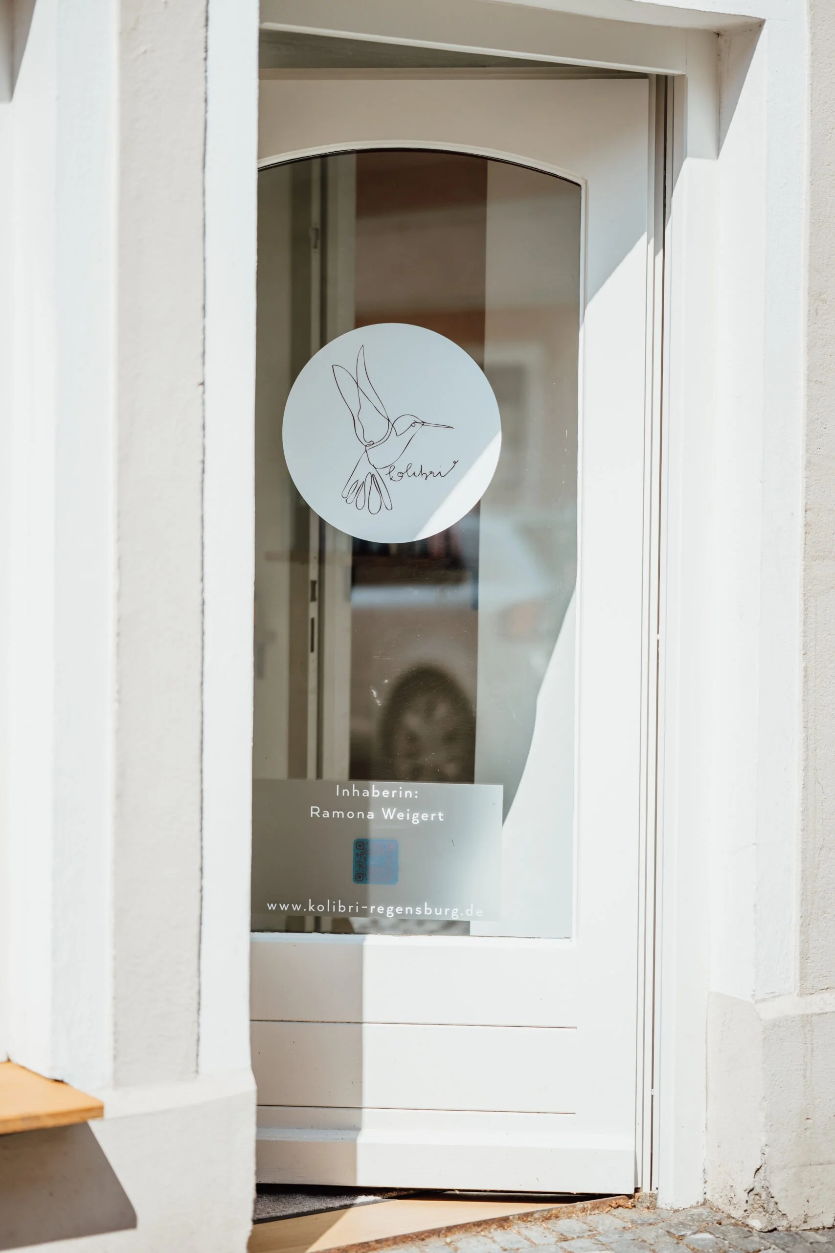 Weiße Tür mit einem Hummel-Logo und einem Schild mit der Aufschrift 'Inhaberin: Ramona Weigert', www.kolibri-regensburg.de, an der Tür. Die Tür ist in einem weißen Rahmen und das Gebäude besteht aus hellen, glatten Oberflächen.