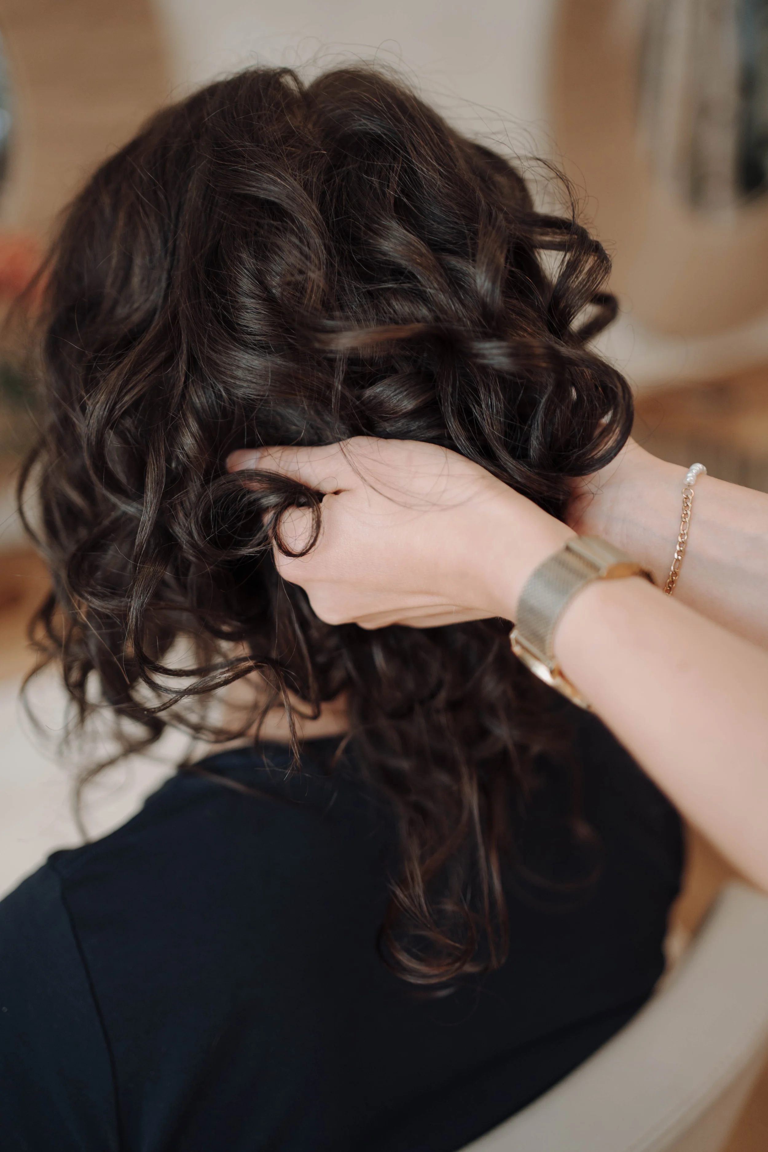 Frau mit lockigem braunem Haar, die sich die Haare am Kopf bindet, trägt eine schwarze Kleidung und Schmuck an beiden Handgelenken.