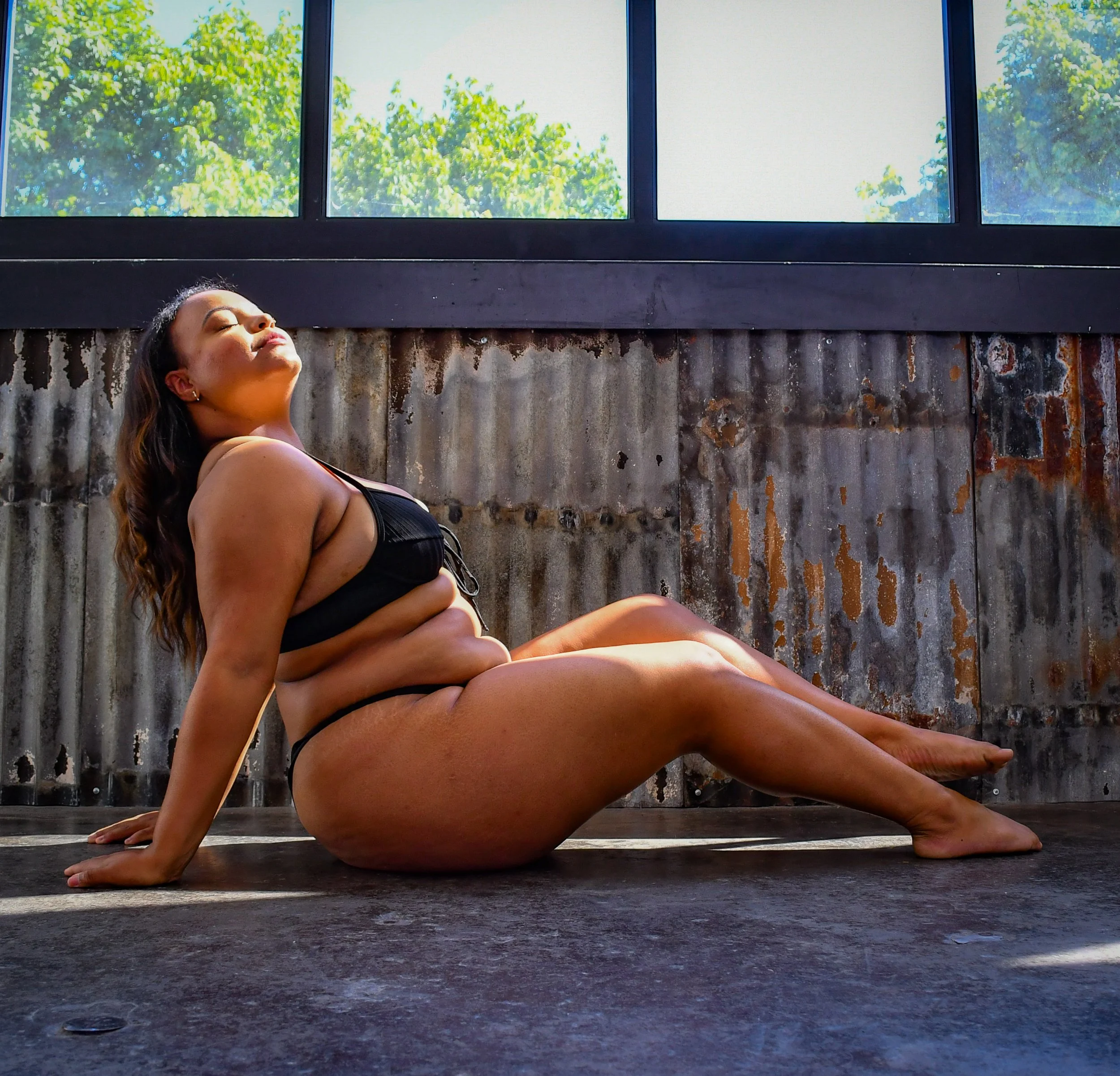 A woman in black lingerie sitting on the floor basking in her natural spray tan for her brown skin with a corrugated metal wall and large windows behind her, with sunlight and green trees outside.