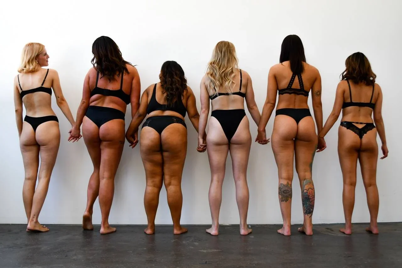 Seven women in underwear showing off their spray tans, holding hands, standing in front of a plain white wall.