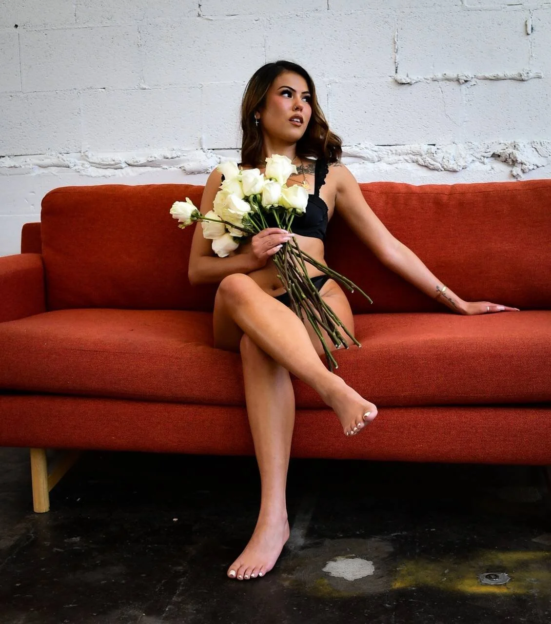 Tan woman with a natural spray tan in black lingerie sitting on a red couch, holding a bouquet of white roses, with a white brick wall background.
