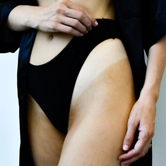 A woman wearing black underwear and a dark robe adjusting her clothing to show off her custom spray tan and natural skin color.