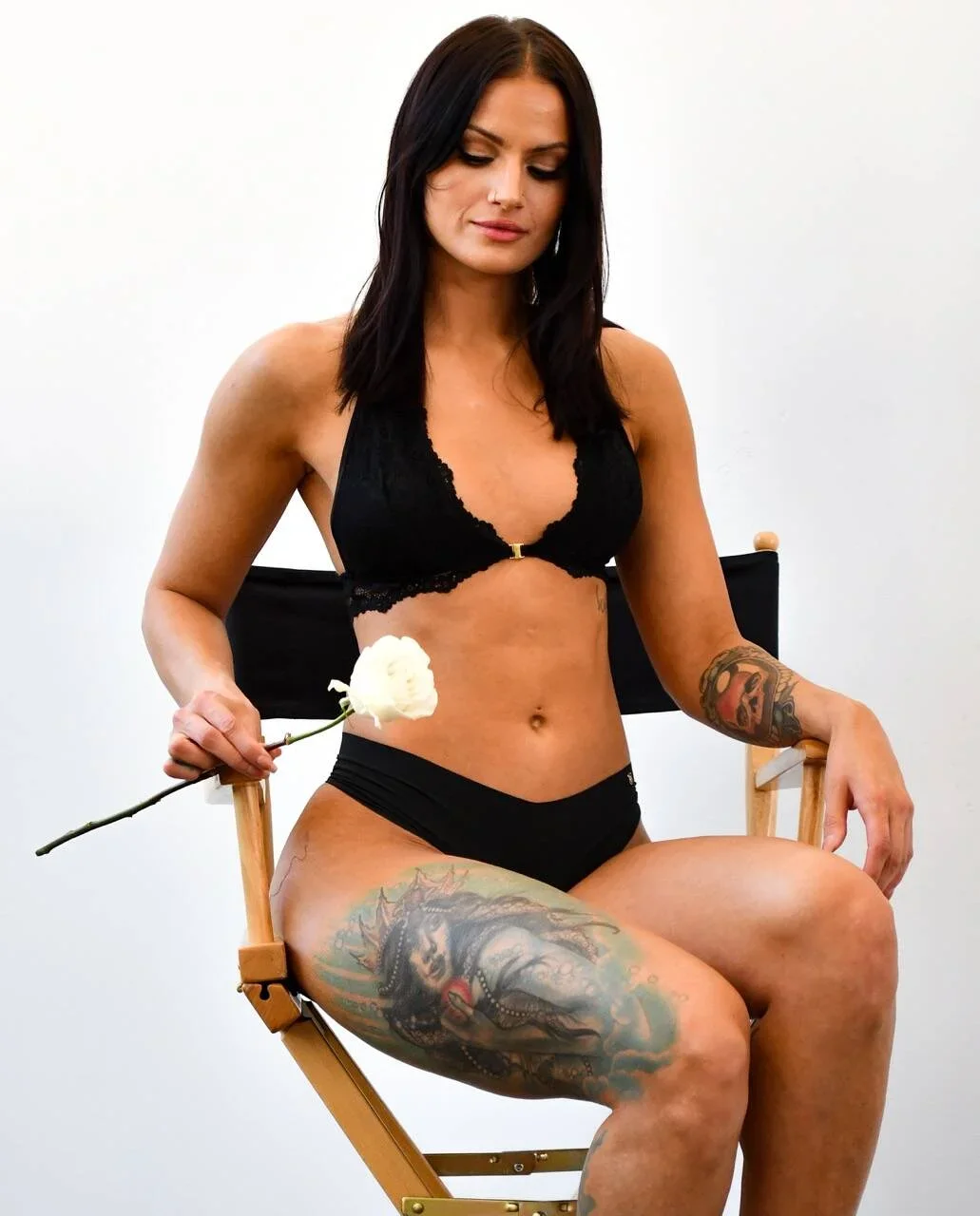 A woman with dark hair sitting on a director's chair, holding a white flower, wearing black lingerie, and displaying a spray tan and tattoos on her thigh and arm against a plain white background.