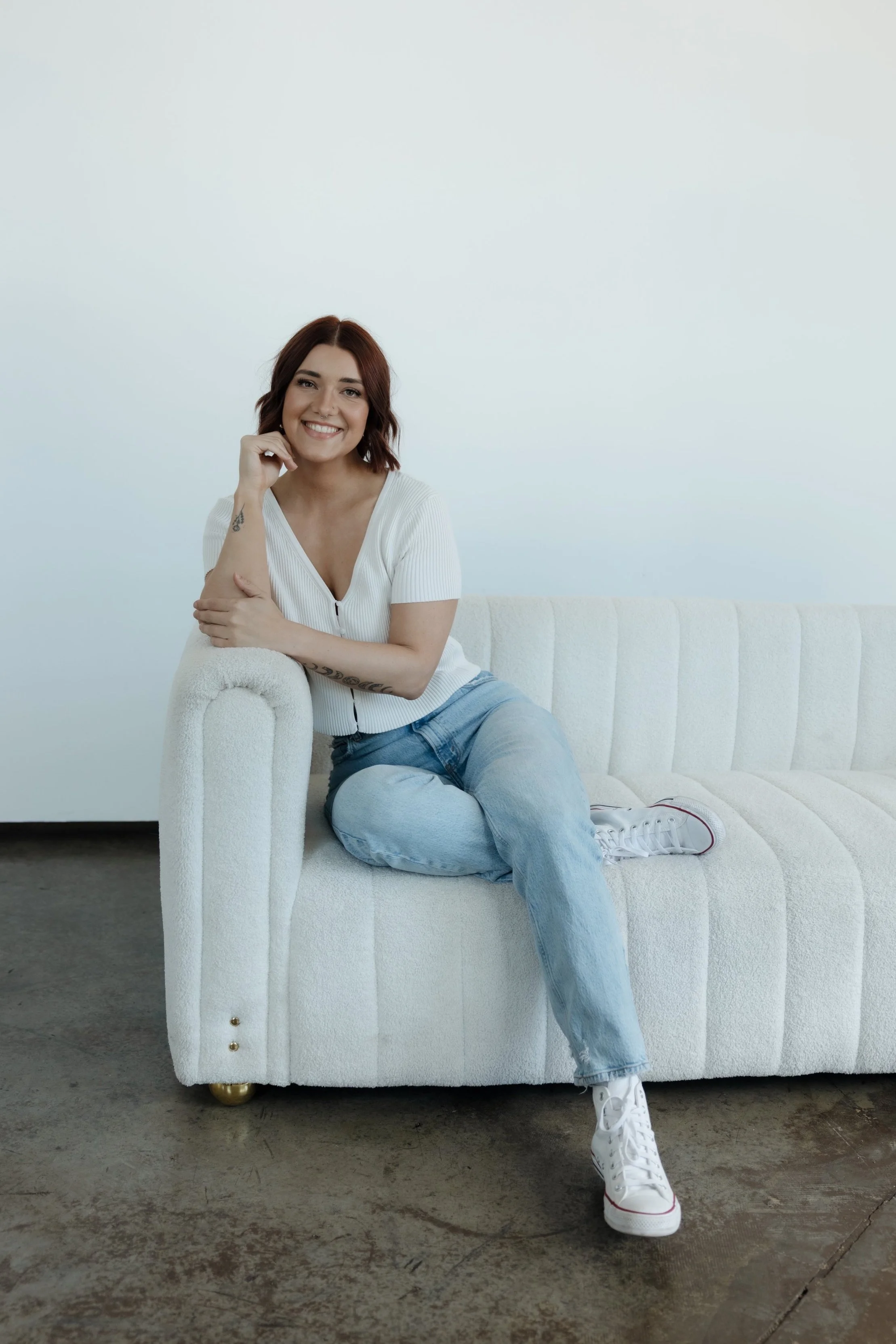A young woman with shoulder-length reddish-brown hair, wearing a white short-sleeved top, light blue jeans, and white sneakers, sitting casually on a white sofa with a plain, light-colored wall in the background. She is smiling and looking at the camera.