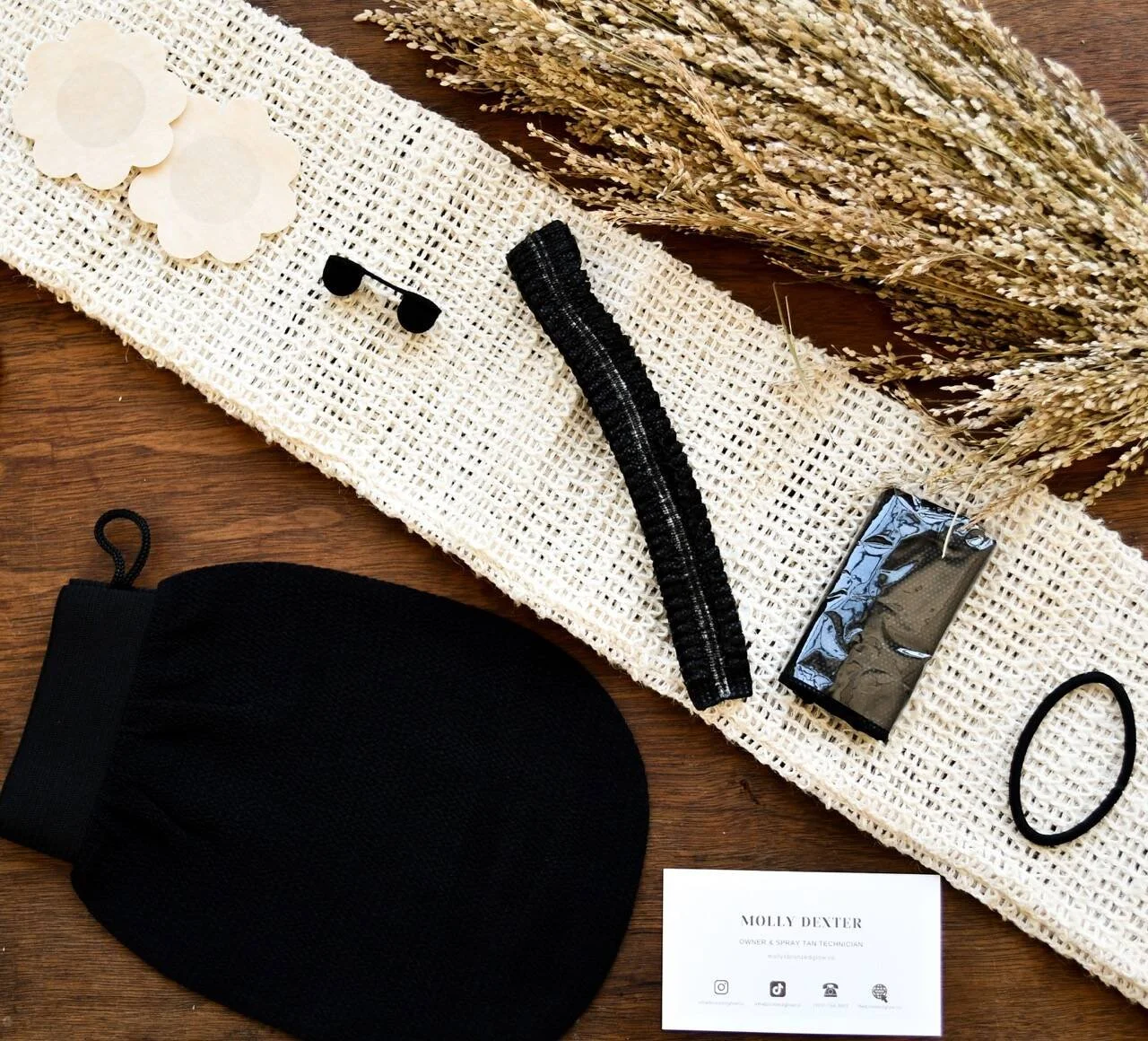 Flat lay of spray tan supplies including, pasties, a nose filter, a hairnet, disposable underwear, a black elastic hair band, an exfoliating mitt and a business card on a wooden surface.