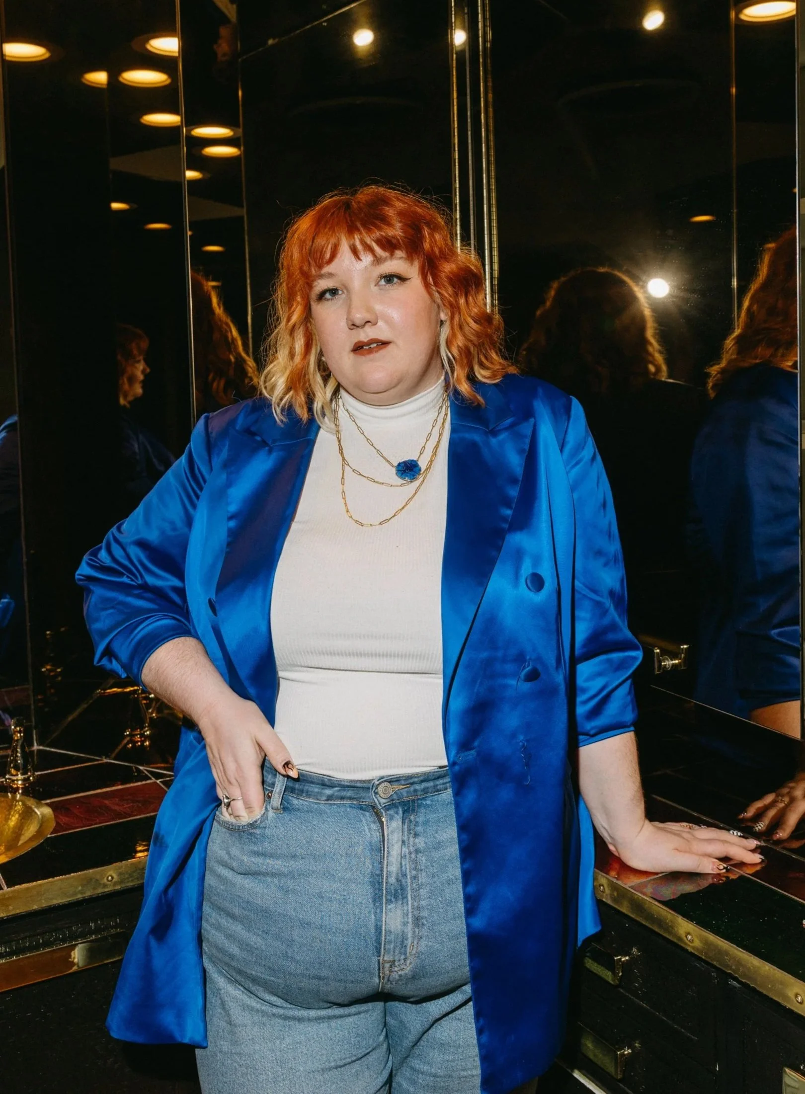 A woman with orange hair wearing a white turtleneck, layered gold necklaces with a prominent blue pendant, and a shiny blue blazer, standing in front of mirrors in a dimly lit, modern bathroom with gold fixtures.