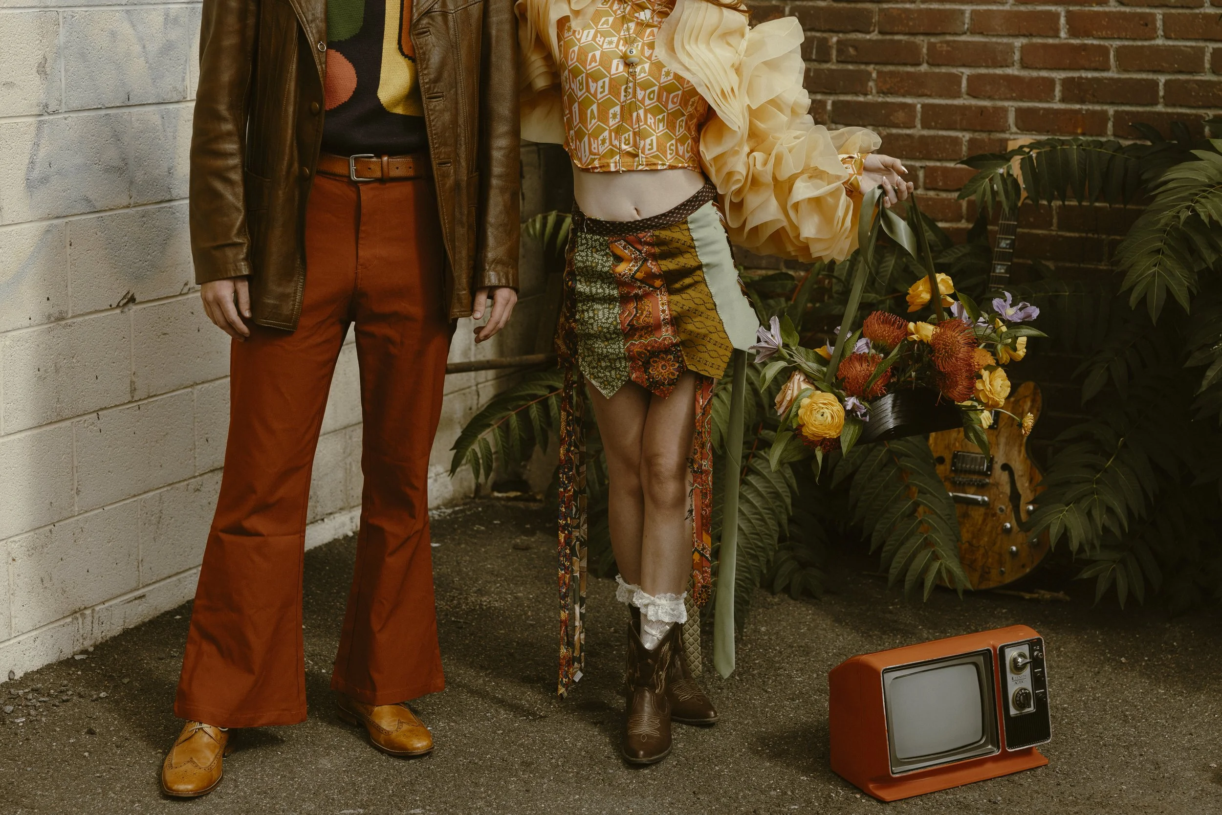 Close-up of two people, one in orange pants and a brown leather jacket, the other in a colorful patchwork skirt and a patterned top, standing in front of a brick wall and plants. On the ground is a vintage orange television set. There are flowers in the background and a guitar partially visible behind the plants.