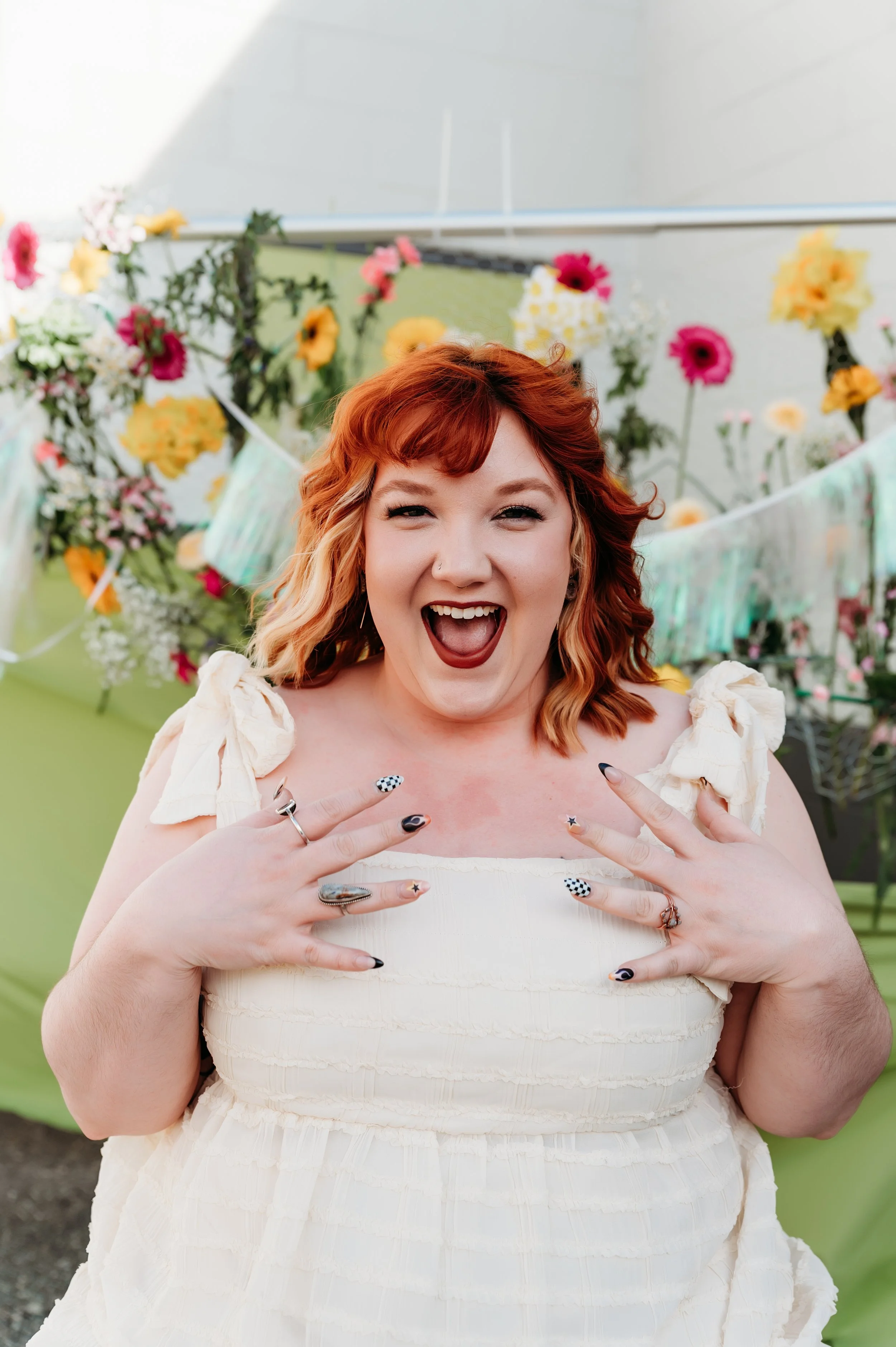 A joyful woman with red hair and dark lipstick smiling with hands near her chest in front of a floral backdrop at a celebration.