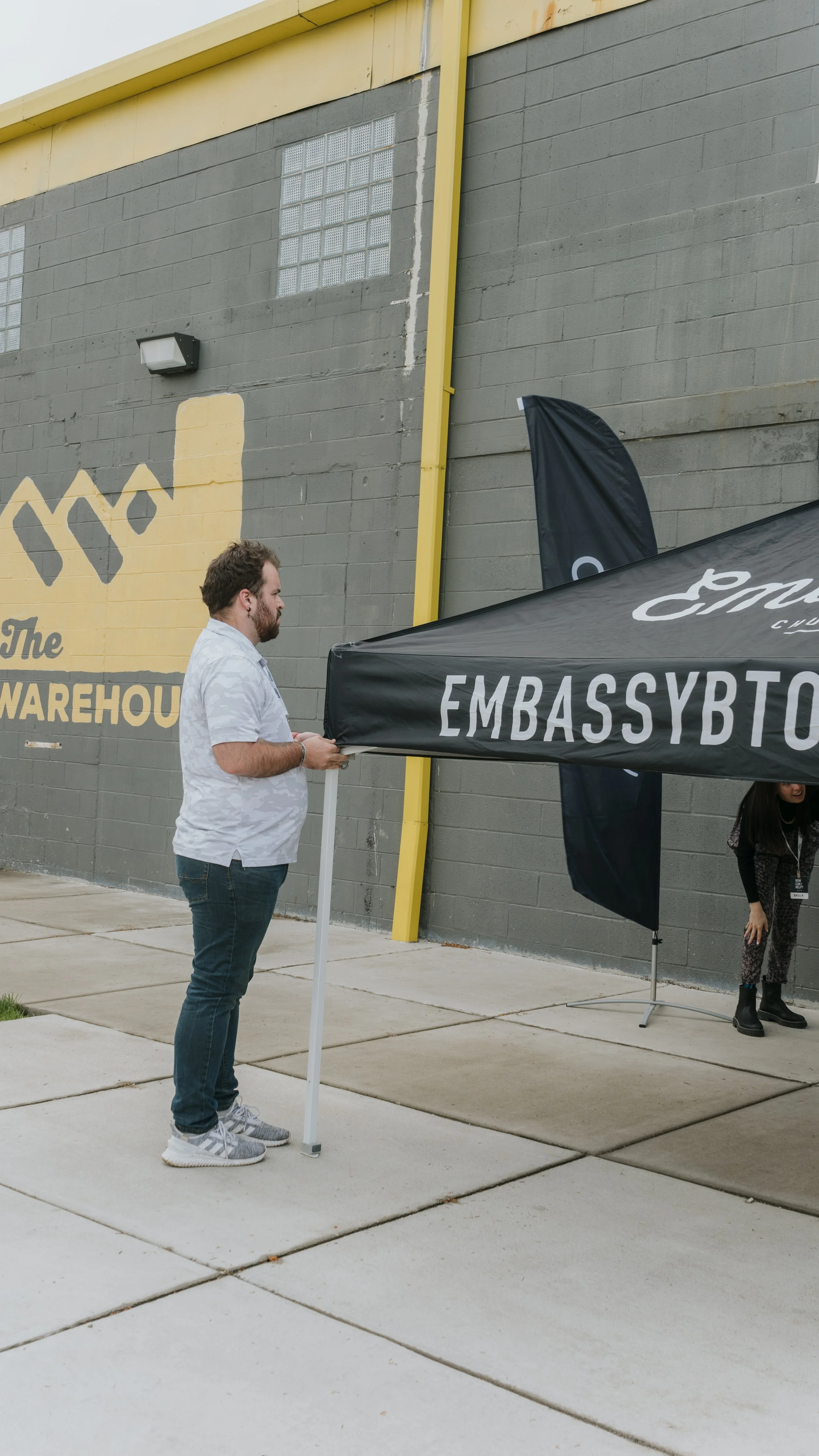A man standing outdoors near a black canopy tent with the word 'EMBASSY' visible, in front of a gray brick wall with yellow accents and a warehouse sign.