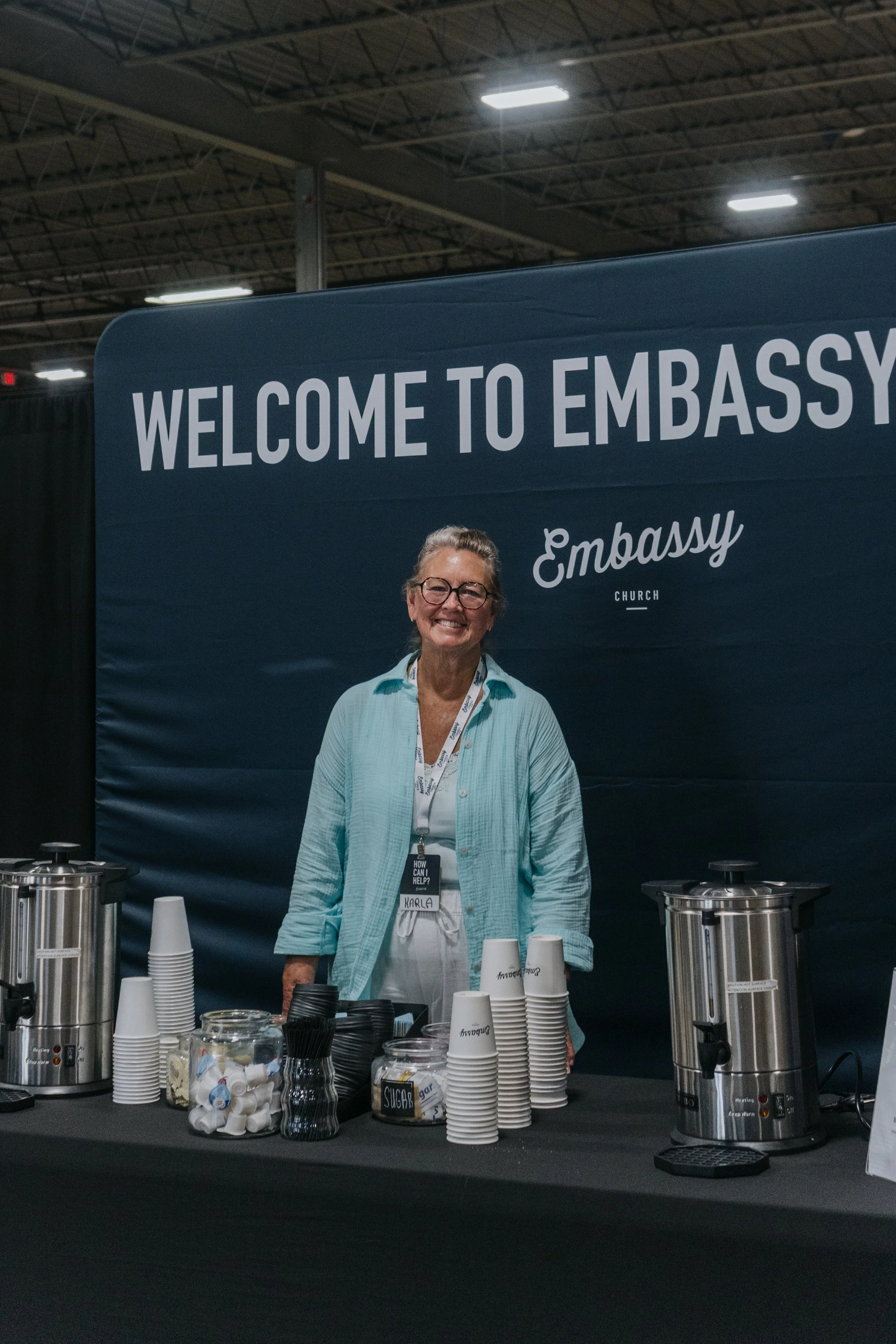 A smiling woman standing behind a coffee station at a welcome desk with a large sign that says 'Welcome to Embassy' in a spacious indoor event space.