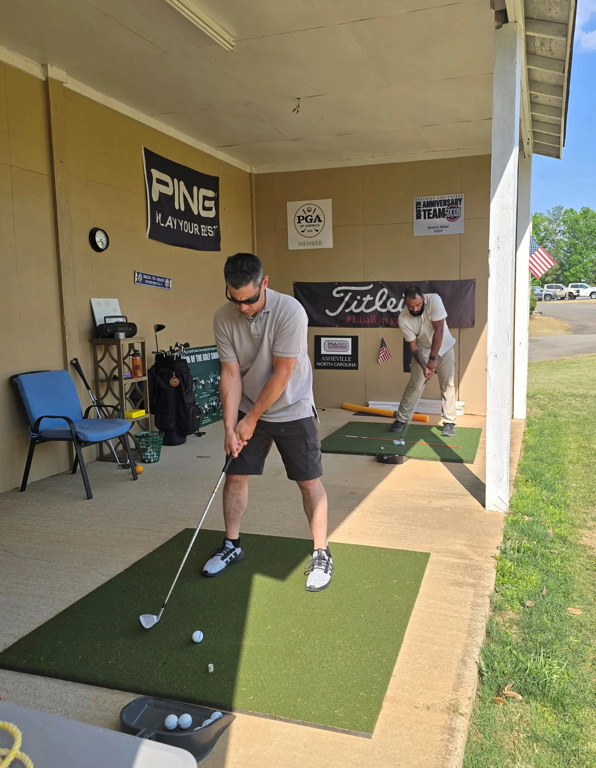 Two people practicing golf swings on indoor mats with golf balls, located in a covered outdoor area with golf banners on the wall.
