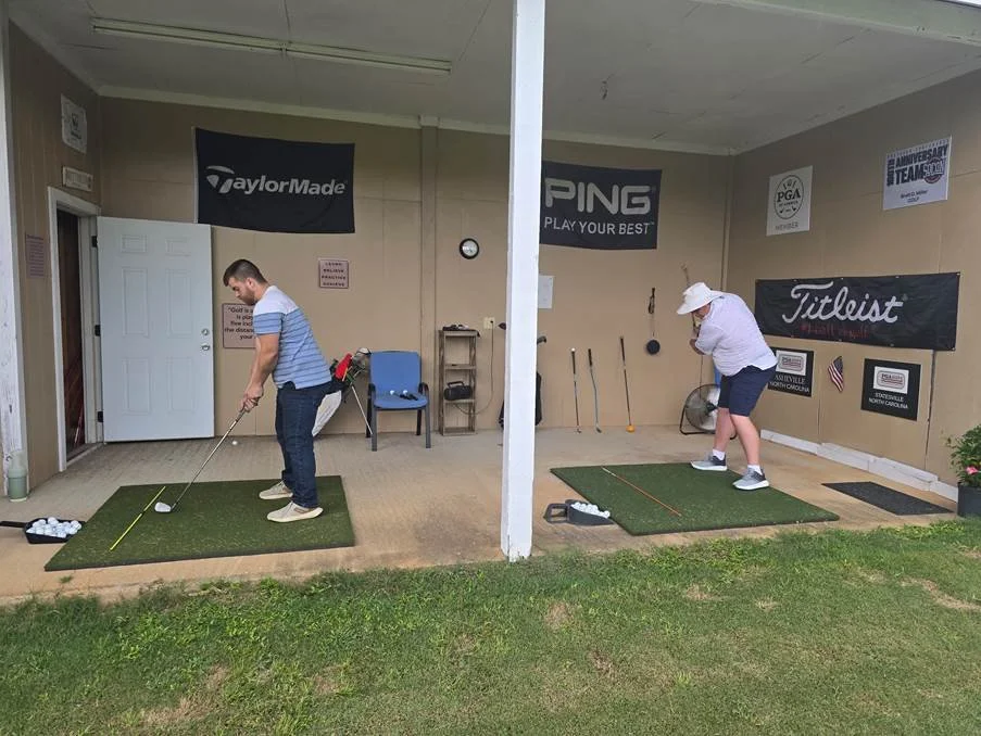 Two people practicing golf swings on indoor mats with golf balls, located in a covered outdoor area with golf banners on the wall.