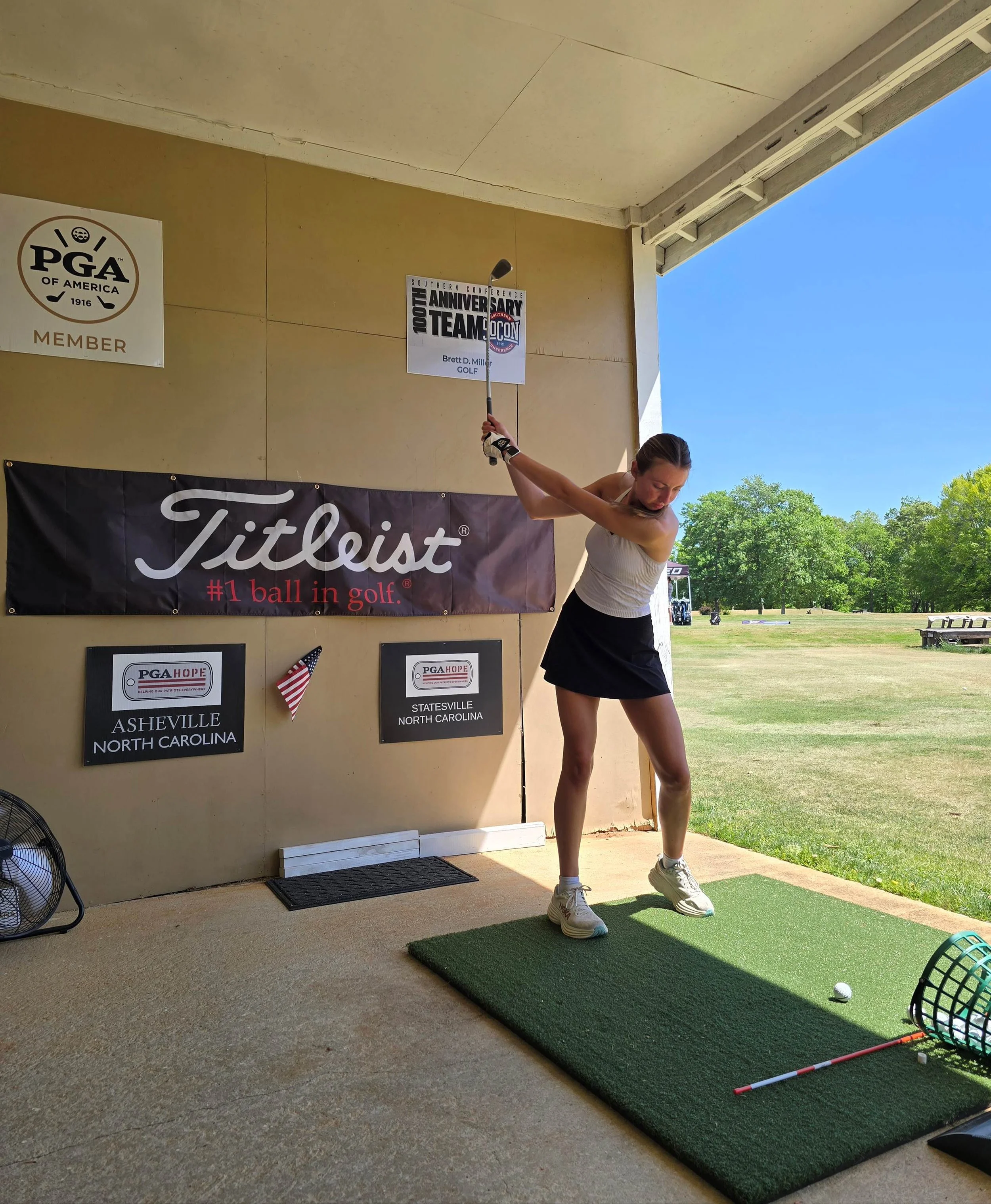 A person teeing off on a golf course with a golf ball on a tee, mid-swing, during daytime with a clear blue sky and trees in the background.