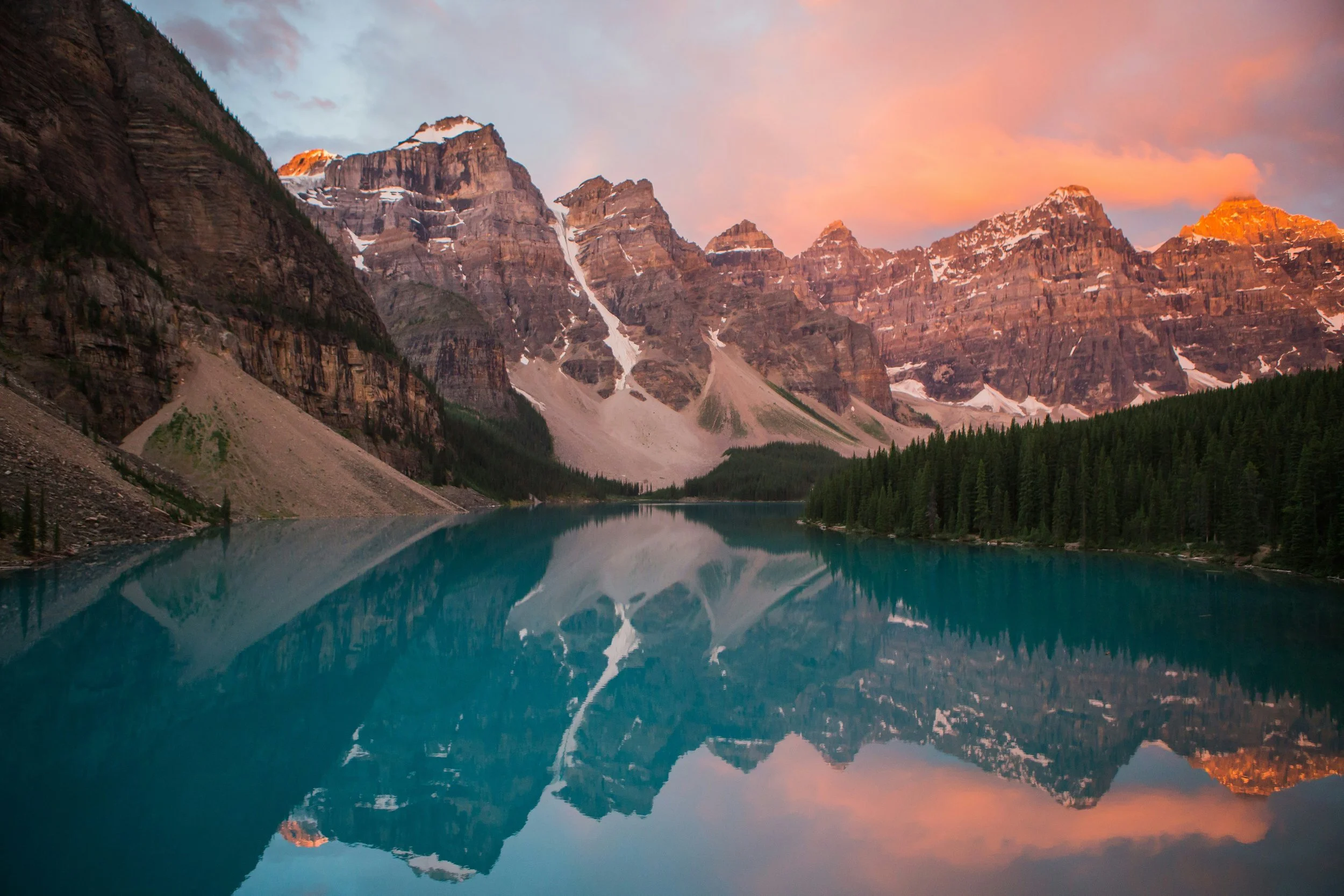 A scenic view of a mountain lake surrounded by tall mountains with snow, and a forest of evergreen trees, with a pink and orange sunset sky.