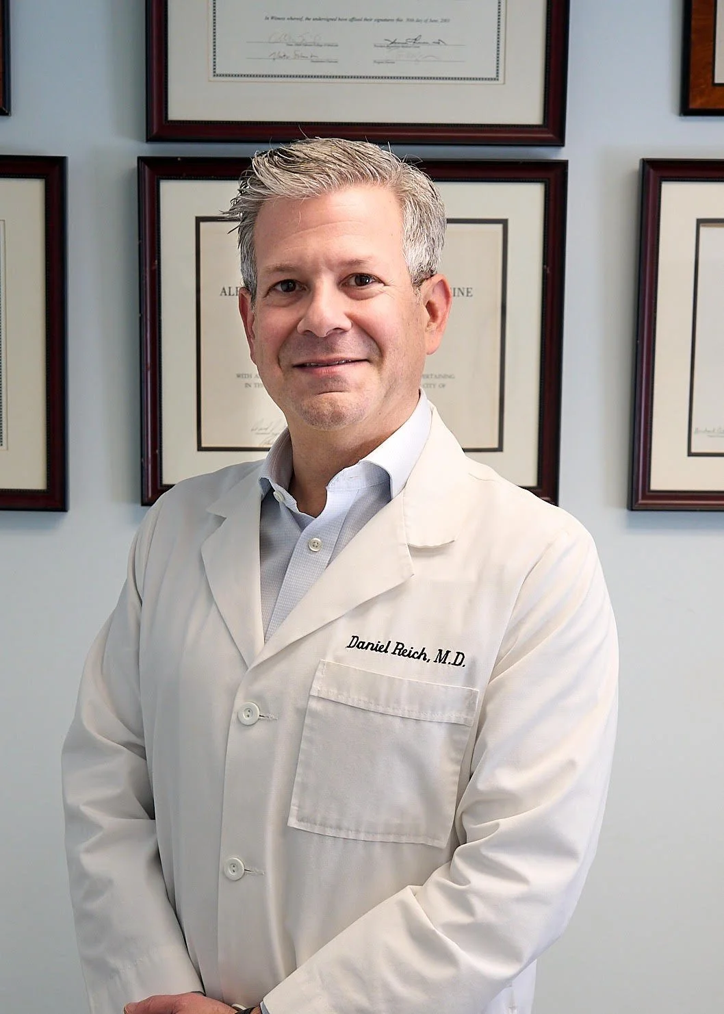 A male doctor wearing a navy suit, orange tie, and a stethoscope around his neck, smiling in a professional setting.