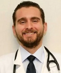 A smiling male doctor with dark hair and beard, wearing a white coat and a stethoscope around his neck.