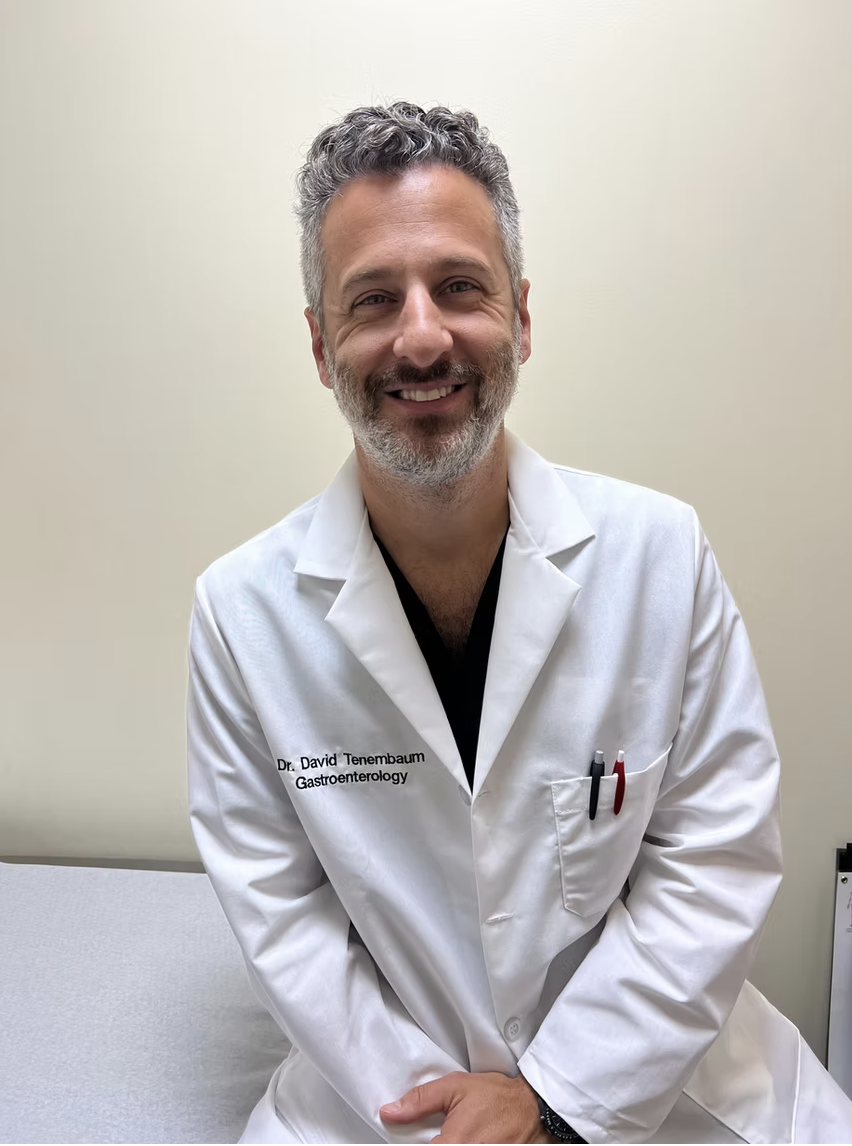 A smiling middle-aged man with curly gray hair and a beard, wearing a white lab coat with the name tag "Dr. David Tenenbaum, Gastroenterology," sitting in a medical office.