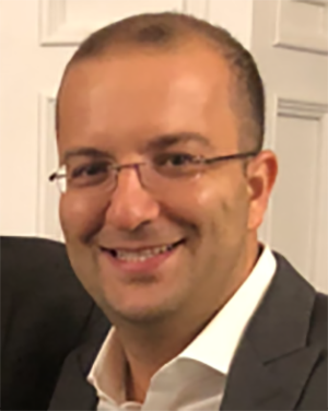 A man with short hair, glasses, and a friendly smile, dressed in a suit, standing in front of a door.