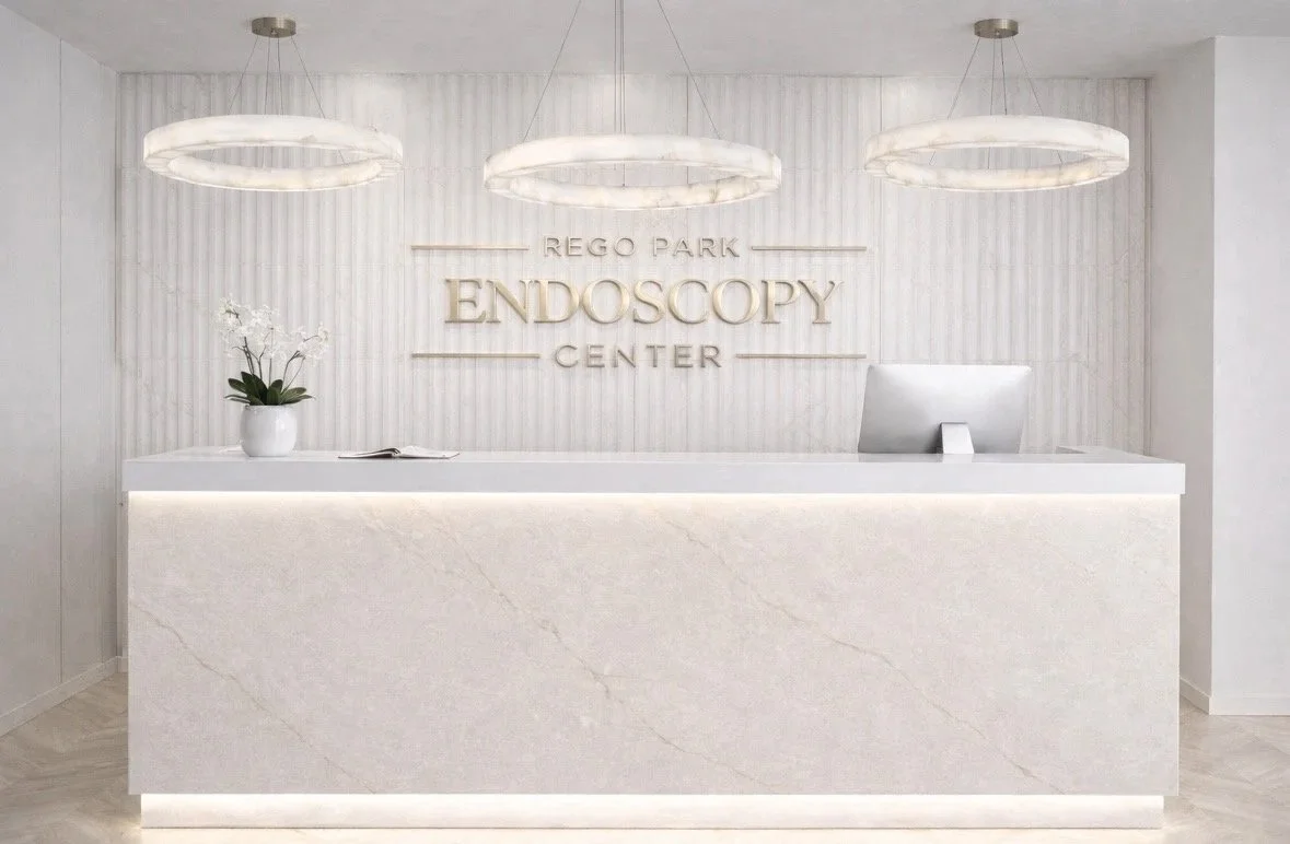 Reception area with a white marble counter and a computer, decorated with a white vase with orchids, and three modern circular ceiling lights. The wall behind displays the sign 'Rego Park Endoscopy Center' in large letters.