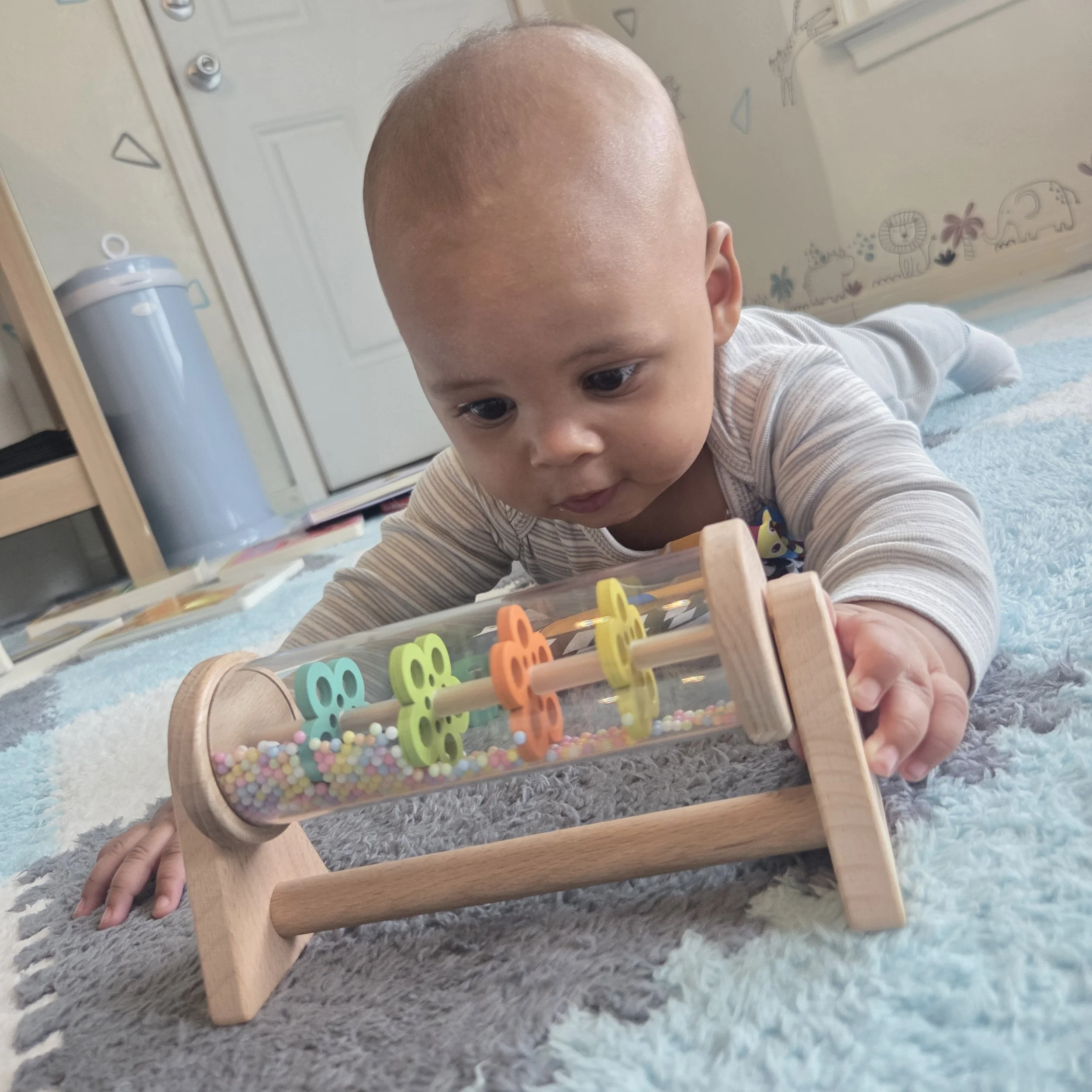Baby in striped long sleeve onesie lying on a blue and white carpet playing with a wooden and glass bead maze toy.