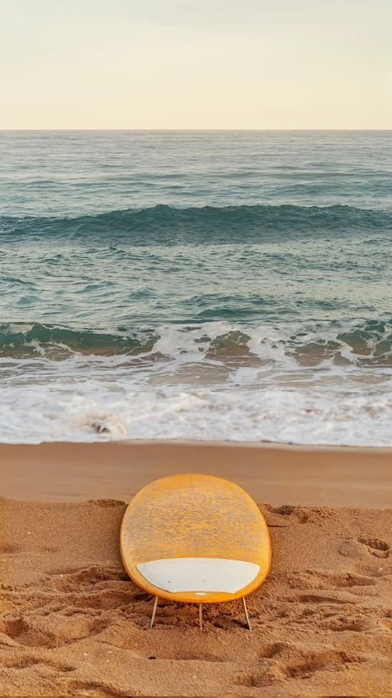 A yellow surfboard resting on the sandy beach with ocean waves and clear sky in the background.