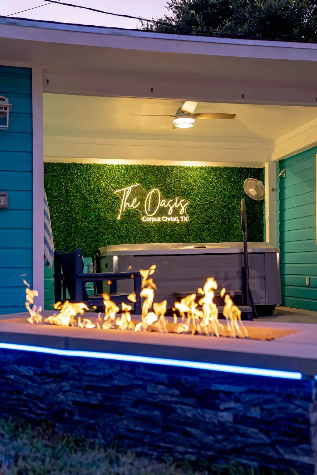 Covered hot tub area with a lit fire pit and 'The Oasis' neon sign at this Corpus Christi vacation rental