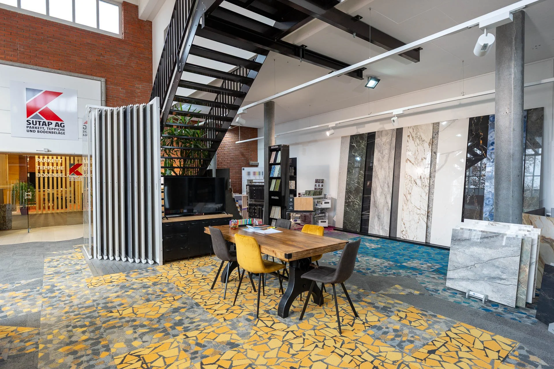 Innenansicht eines Geschäfts für Bodenbeläge mit Stufen, Tischen, Stühlen, Fliesenmustern und Mustertafeln an einer Wand.