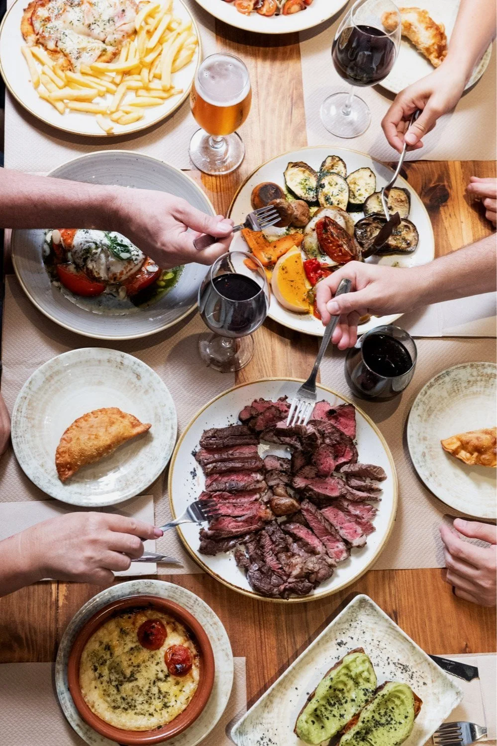 Mesa con diversas comidas, incluyendo carne asada, verduras asadas, milanesa napolitana, ensalada, vino, y otros aperitivos, con varias manos sirviéndose y compartiendo la comida.
