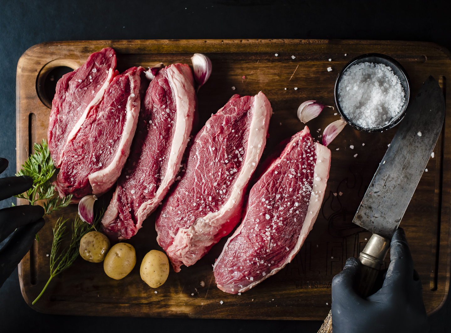 Cinco filetes gruesos de carne de res en un tabla de madera, con sal, ajo, papas pequeñas, hierbas, y un cuchillo, lista para cocinar.