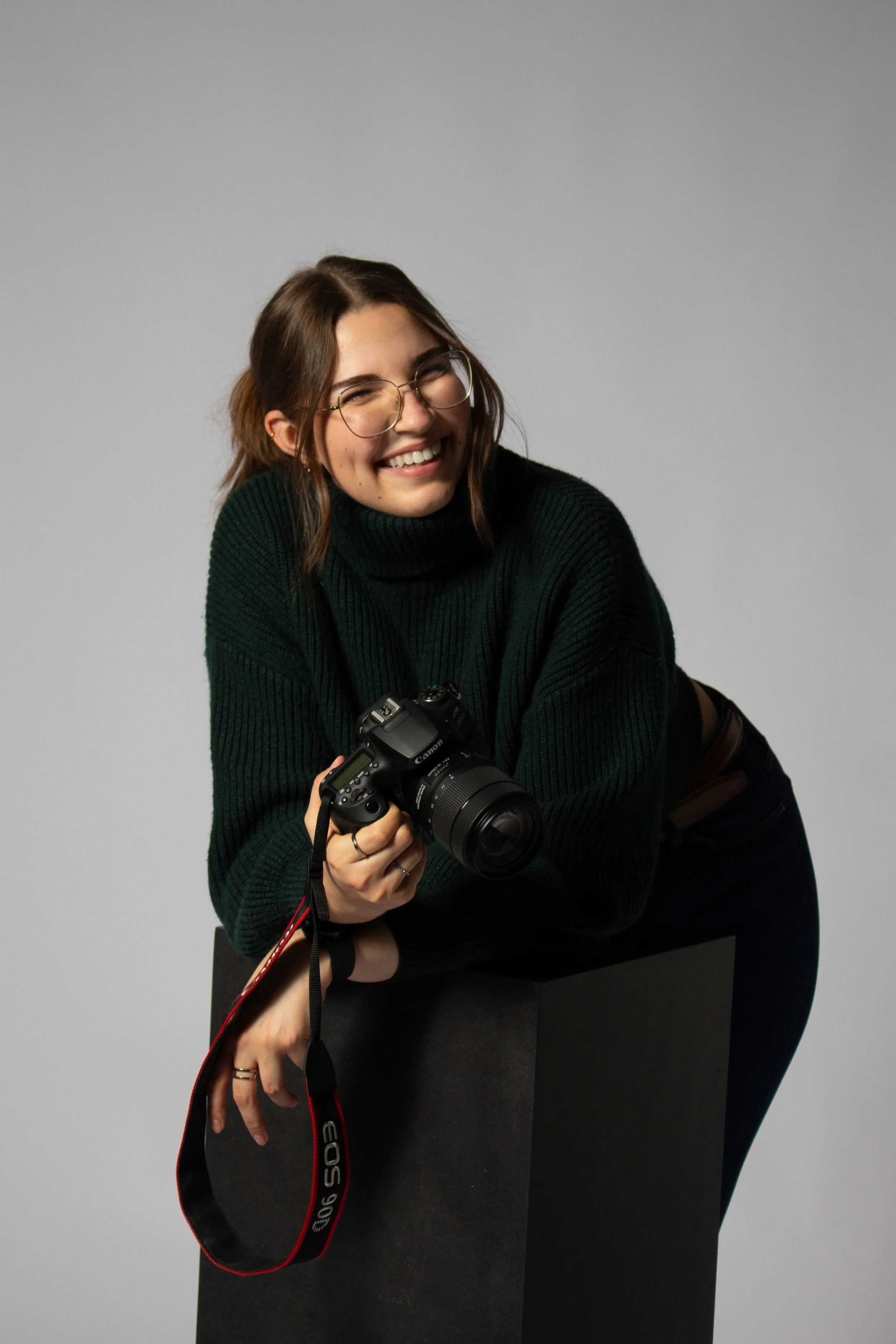 Lachende junge Frau mit Brille, dunklem Pullover, hält Kamera, lehnt sich über schwarzen Block, heller neutraler Hintergrund.