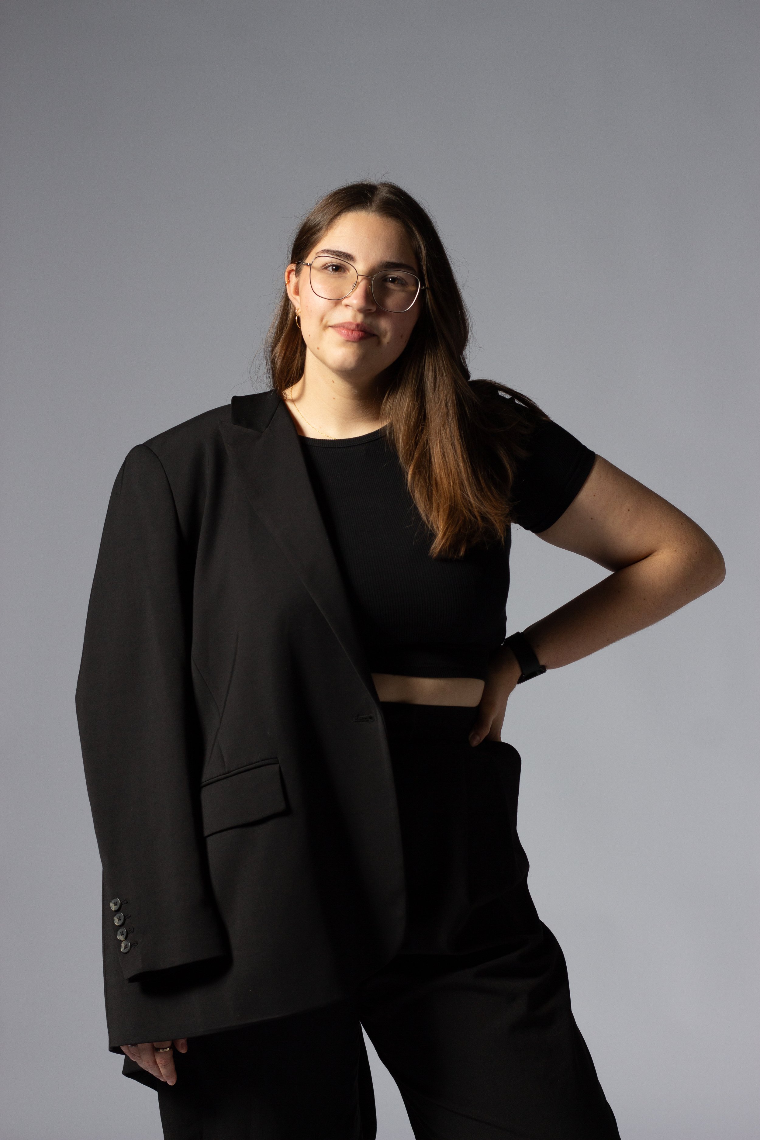 Junge Frau mit Brille, schwarzen Kleid und Anzugjacke vor grauem Hintergrund.