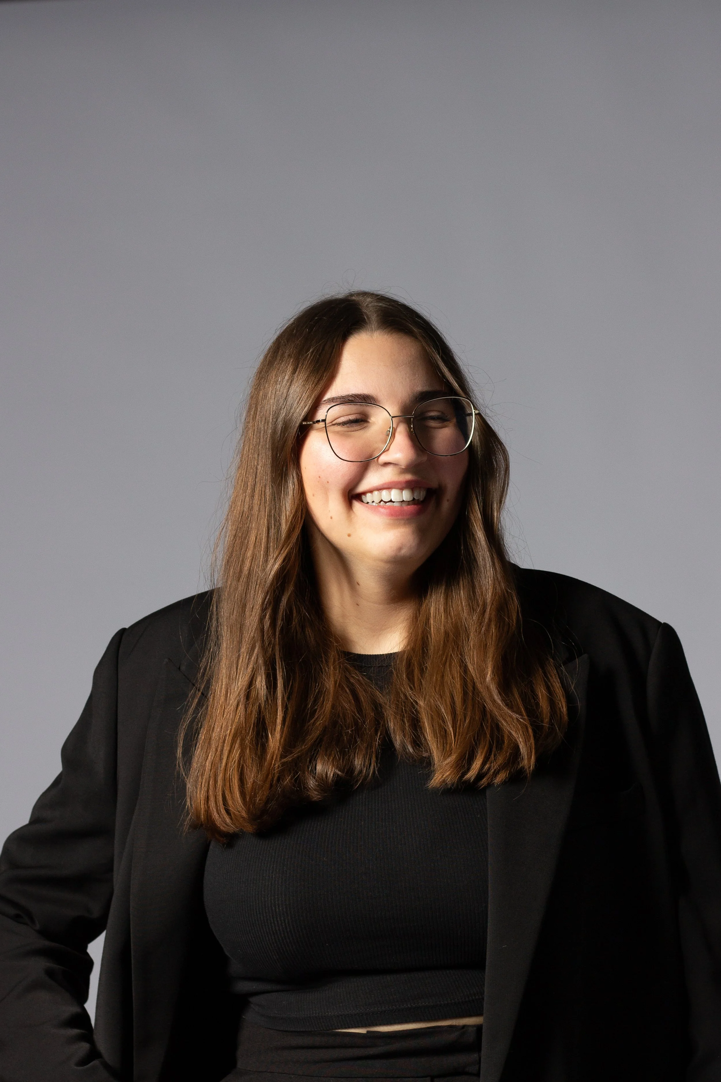 Lächelnde junge Frau mit Brille, schwarze Jacke und schwarzes Top, auf grauem Hintergrund.