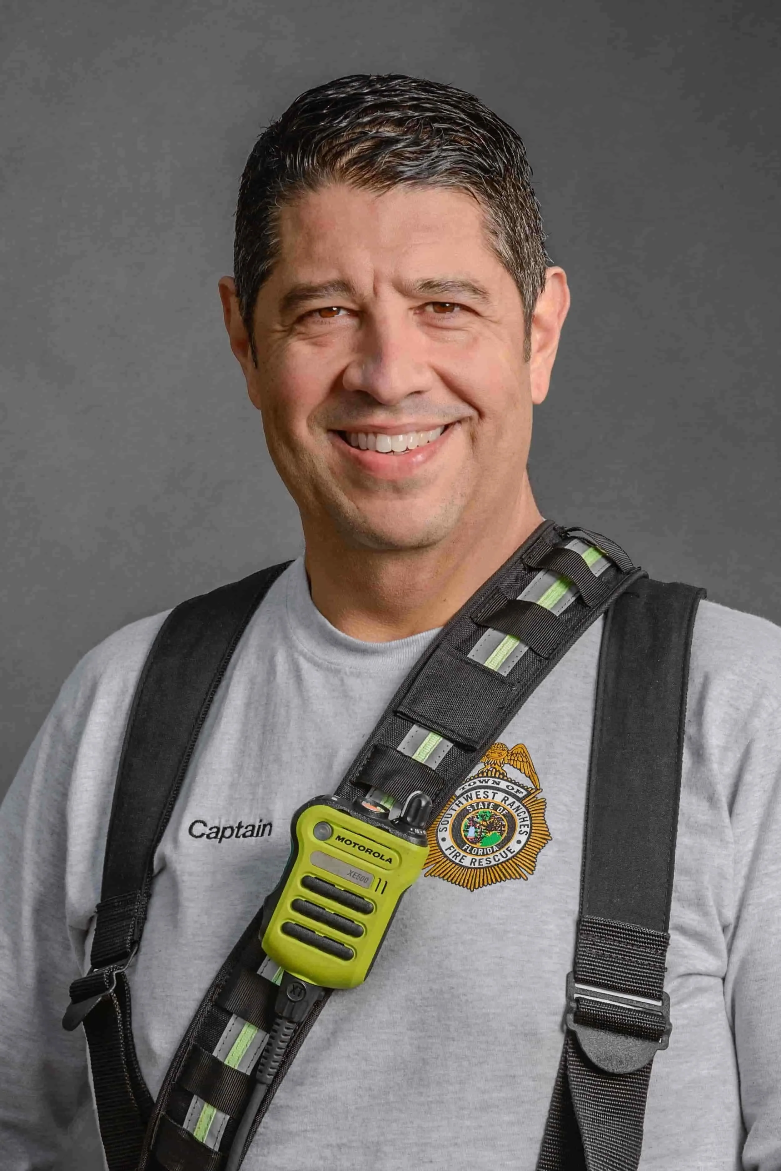 A smiling man in a gray shirt with a fire rescue badge, wearing a shoulder strap with a Motorola radio.
