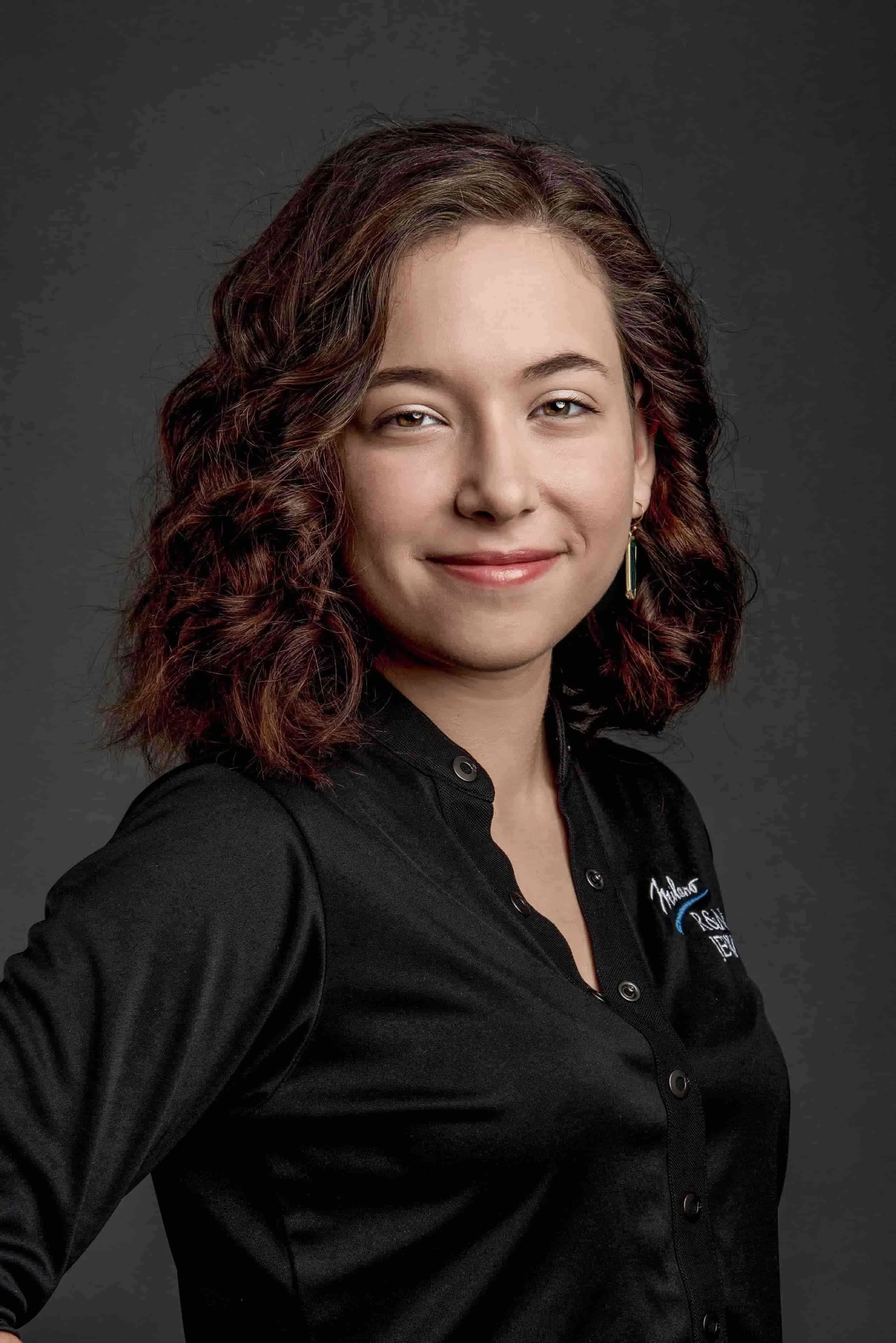 A young woman with shoulder-length curly hair, wearing a black collared shirt with embroidered logo, smiling slightly against a dark gray background.