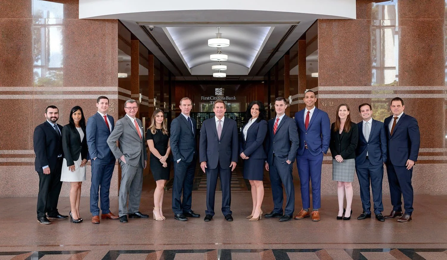 A group of business professionals in formal attire standing in a line inside a bank lobby.