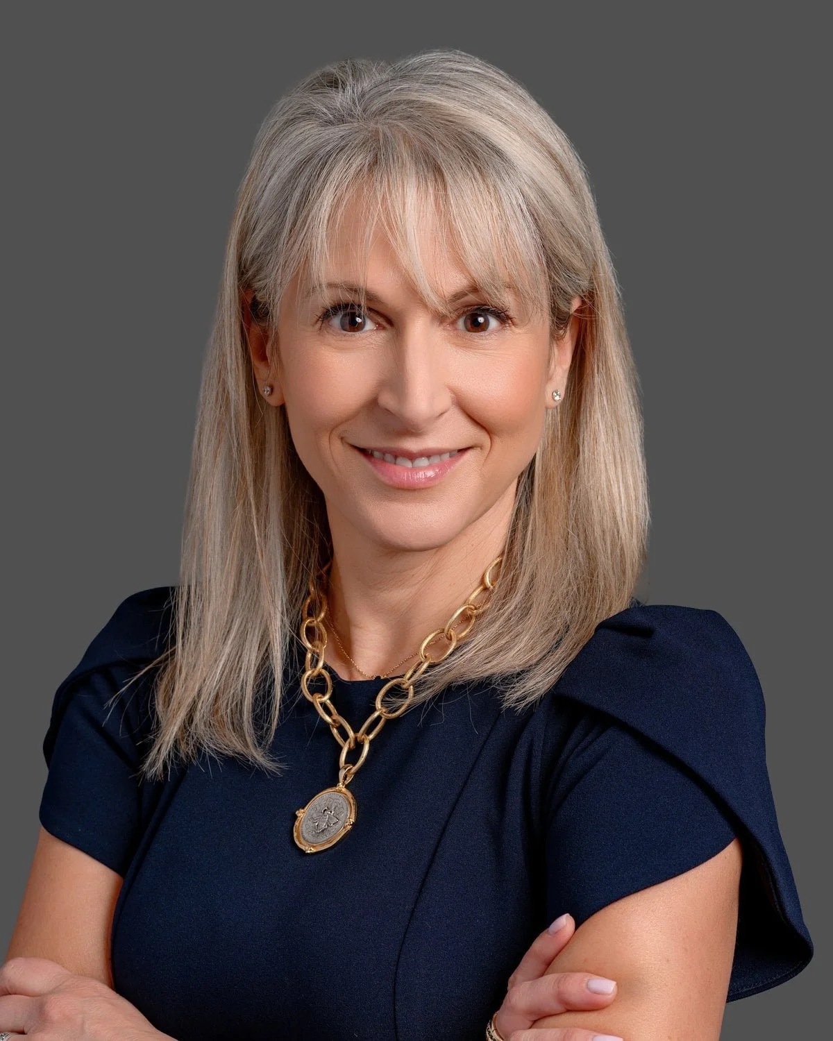 A professional woman with blonde hair, wearing a navy blue dress, gold chain necklace, and small earrings, smiling against a gray background.