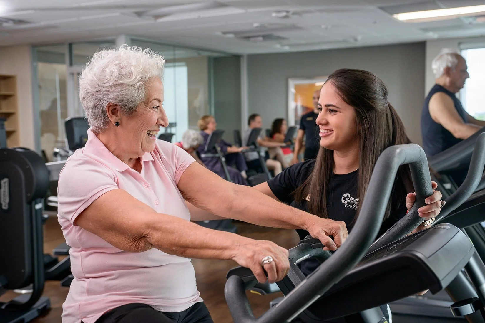 An elderly woman on a treadmill exercising and smiling, with a young female trainer assisting her, in a gym setting with other people exercising in the background.