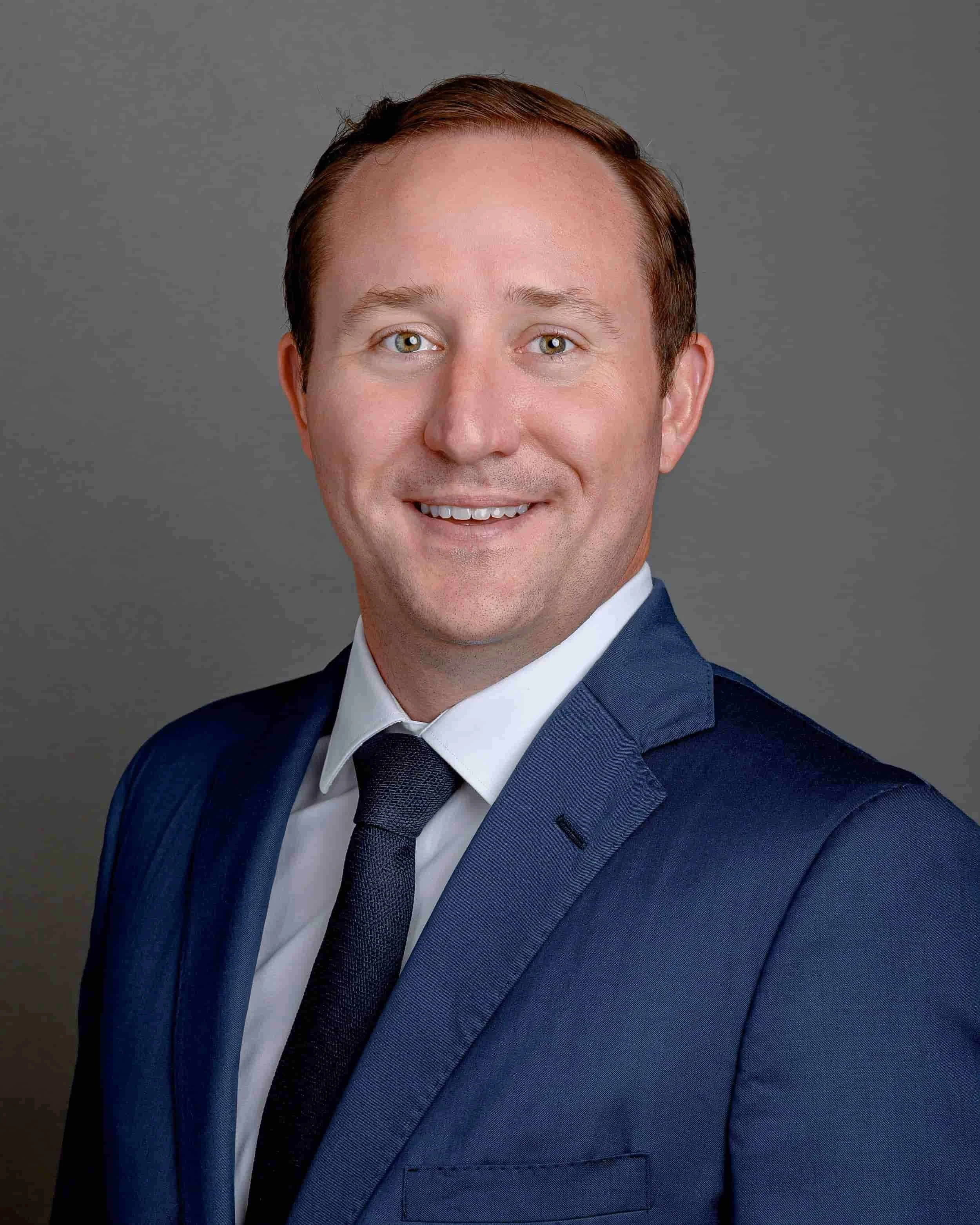 Headshot of a man with light skin, light brown hair, and blue eyes, wearing a dark blue suit, white shirt, and a dark tie, against a gray background.