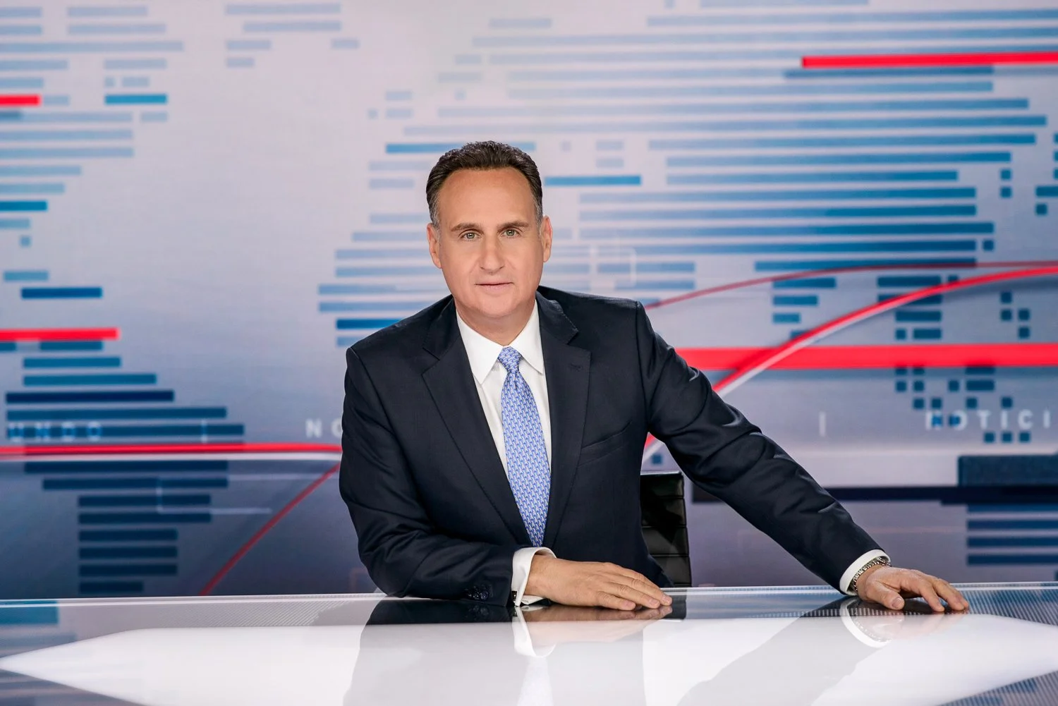 News anchor sitting at a desk in a television studio with a digital background.