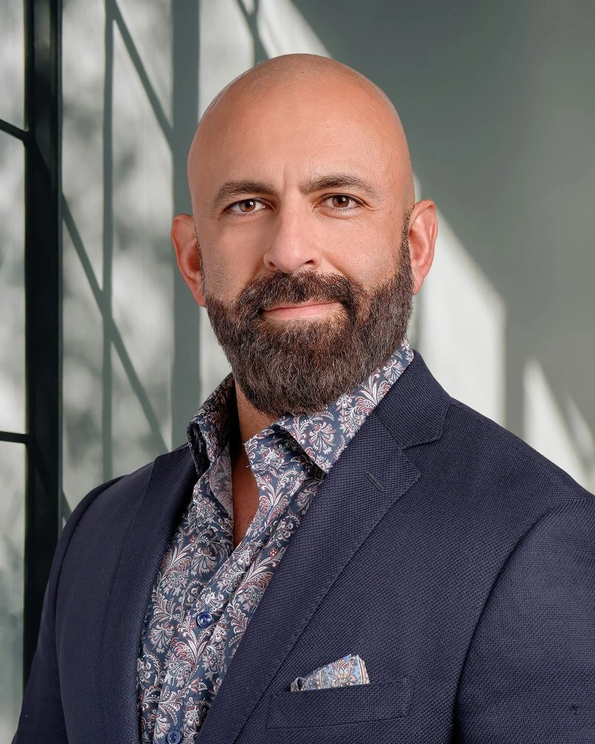 A professional man with a beard and a shaved head, wearing a navy blazer and a patterned shirt, standing in front of a modern glass building.