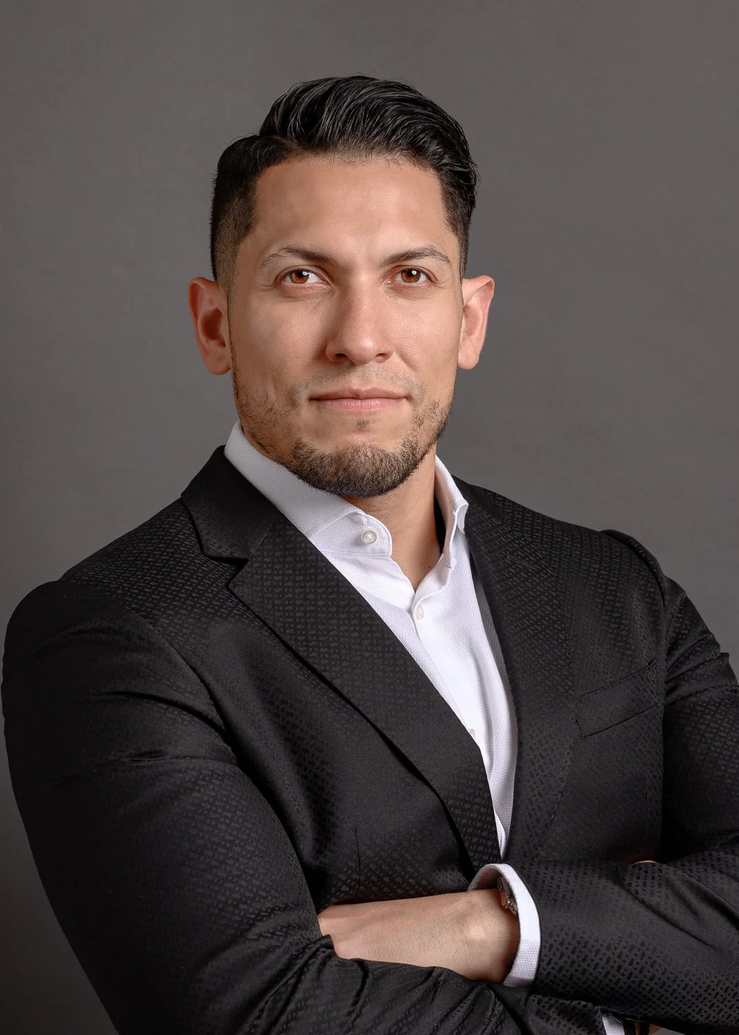 Professional headshot of a man with dark hair, beard, wearing a black suit and white shirt, posing with arms crossed against a gray background.