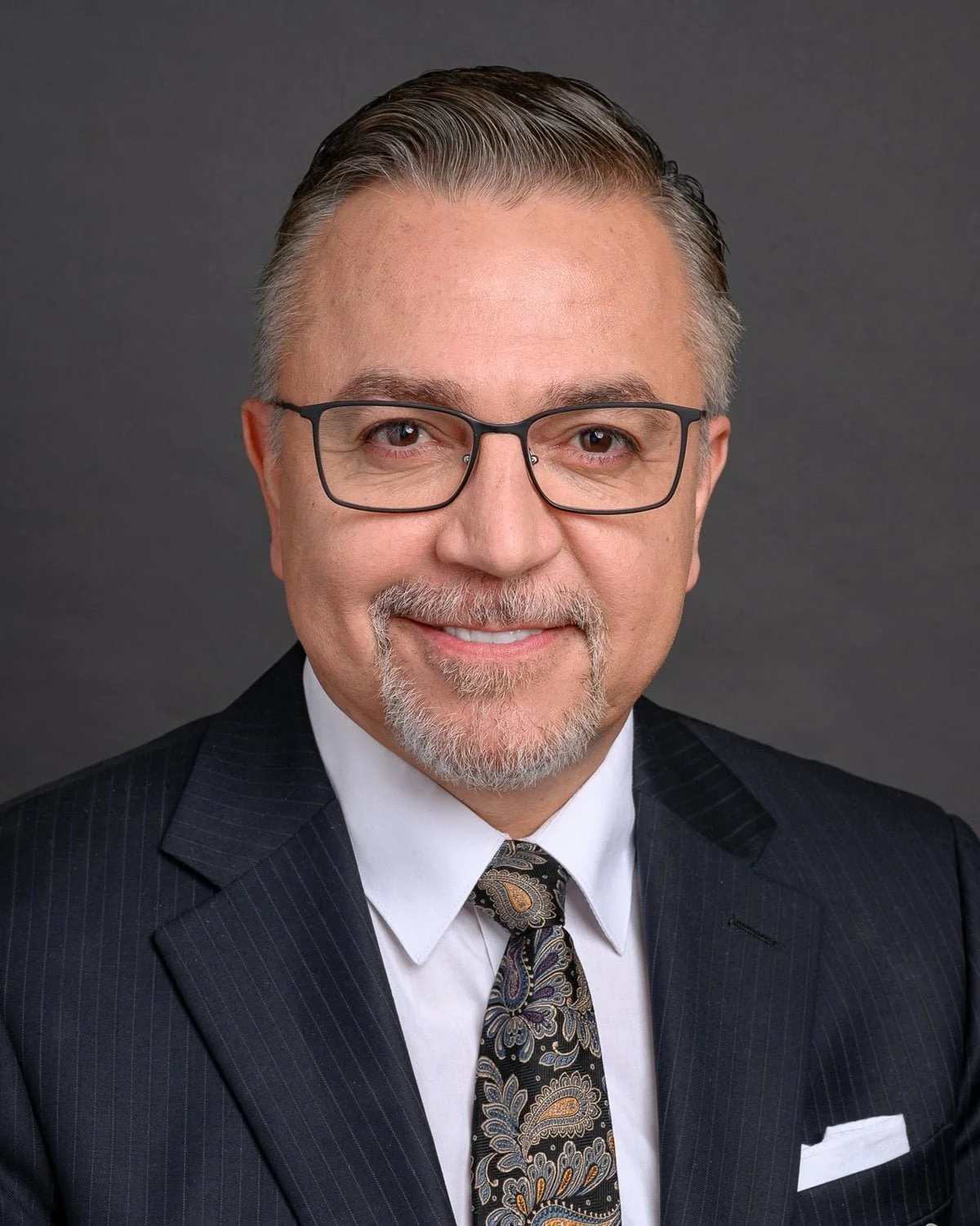 A professional headshot of a middle-aged man with gray hair, glasses, and a goatee, wearing a dark suit, white shirt, and patterned tie, standing against a gray background.