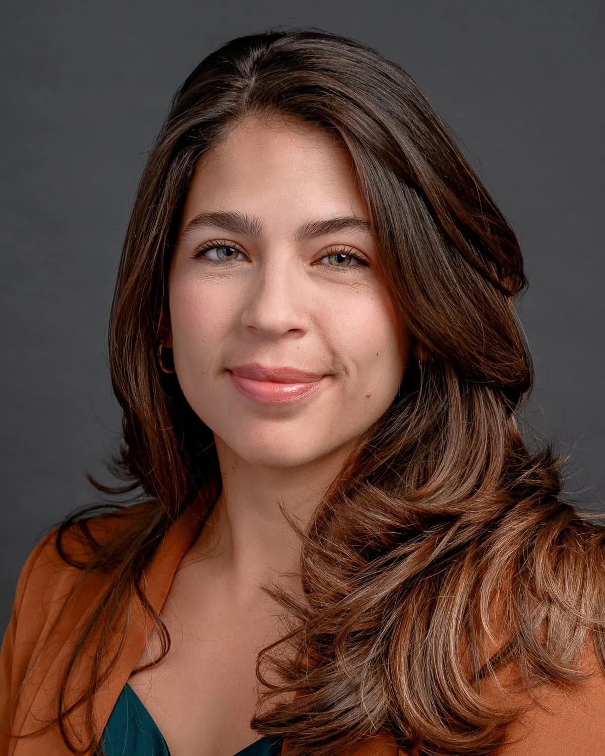 A woman with long brown hair, blue eyes, and light makeup smiling at the camera against a dark gray background.