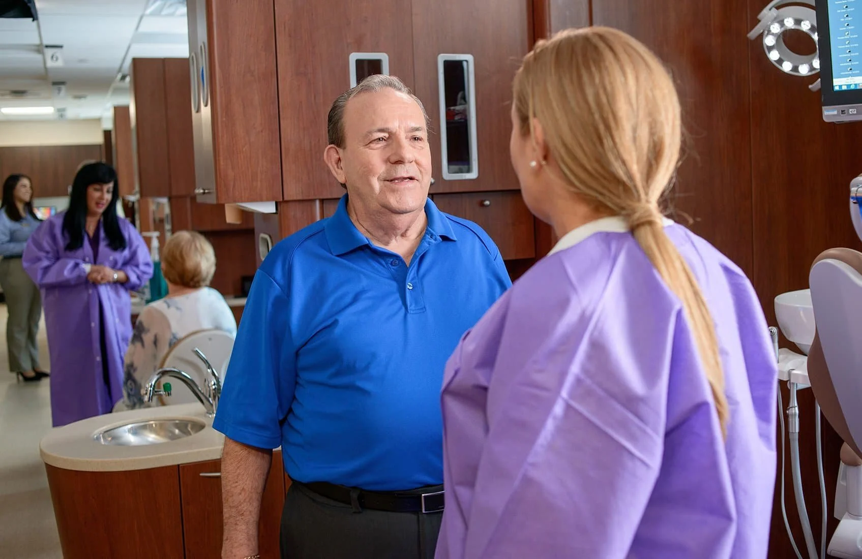 A man and a woman in medical scrubs having a conversation in a healthcare setting, with other healthcare workers in the background.