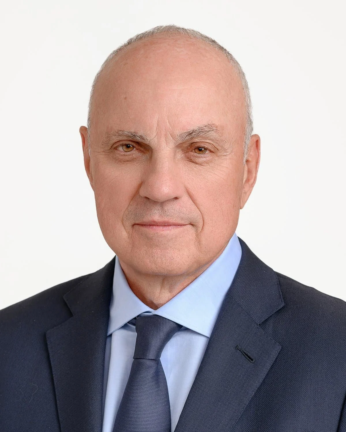 Portrait of an older man with a shaved head, wearing a navy suit, light blue shirt, and navy tie, against a white background.