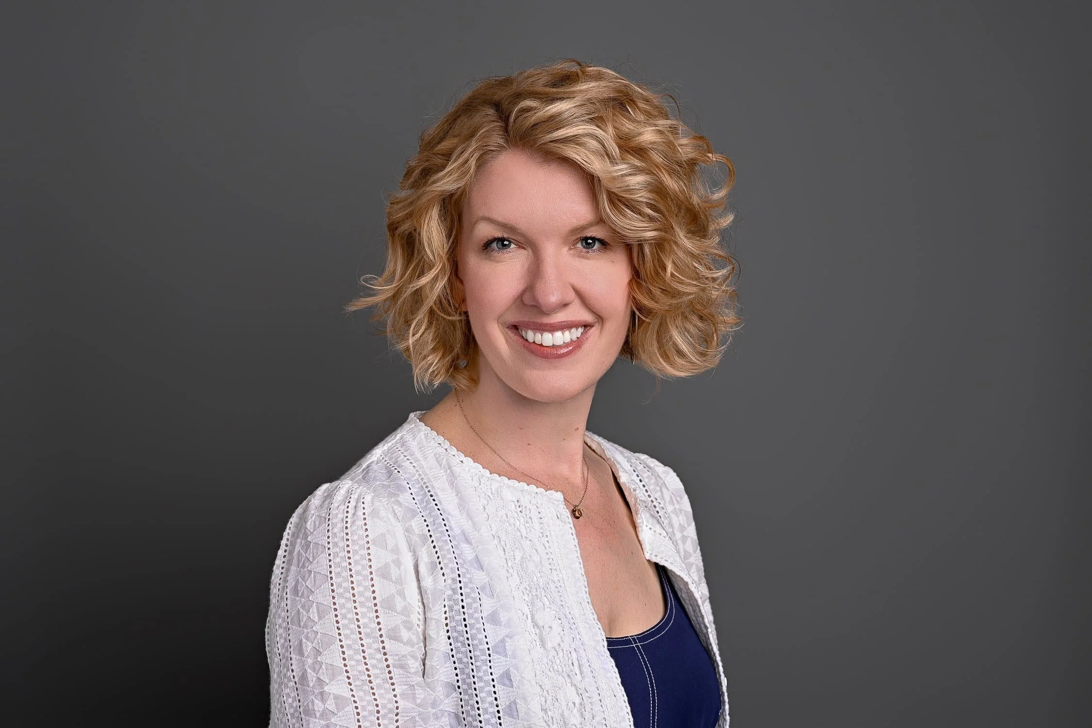 Portrait of a smiling woman with curly blonde hair wearing a white lace top and a navy blue shirt, standing against a dark gray background.