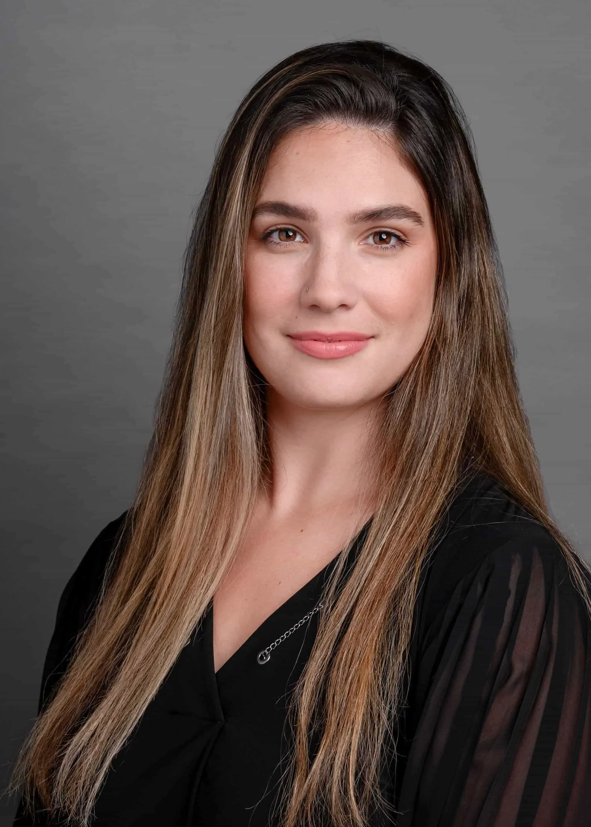 Portrait of a young woman with long brown hair wearing a black top and a silver necklace, smiling softly against a gray background.